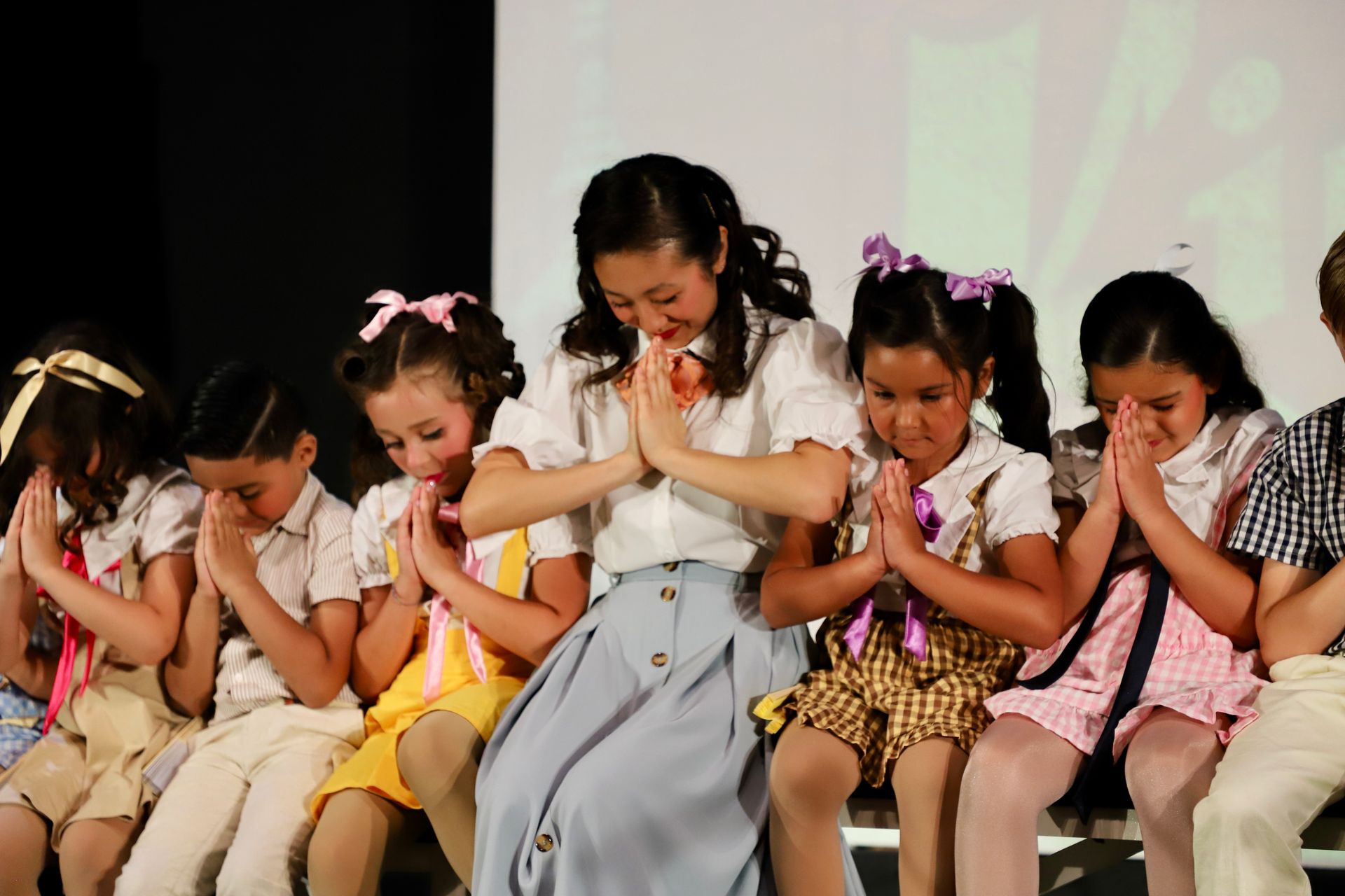 Six children sit on stage, heads bowed and hands together in a synchronized dance pose for a theater performance.