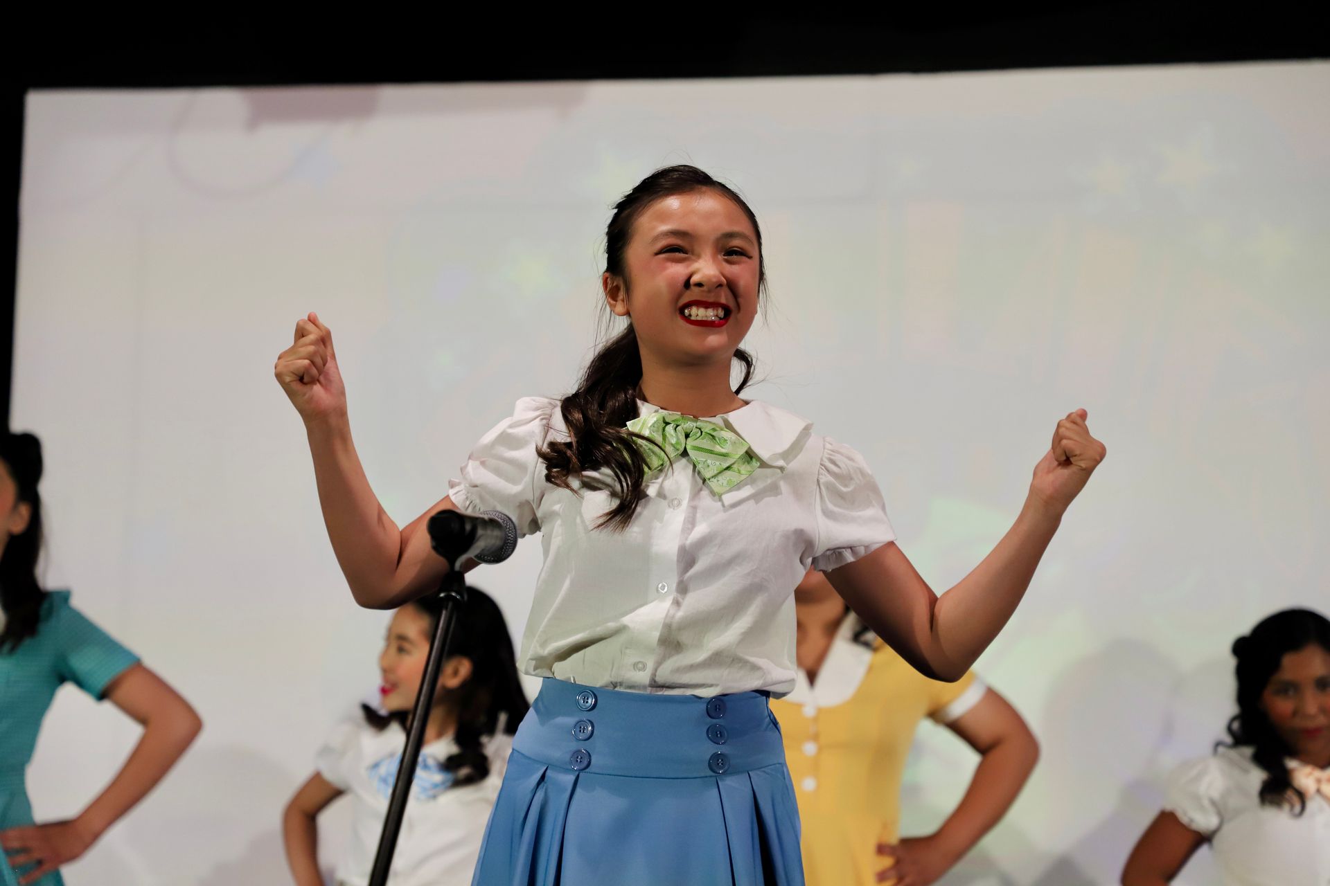 Woman singing or dancing onstage with raised fists, wearing a white blouse and blue skirt for a theater performance.