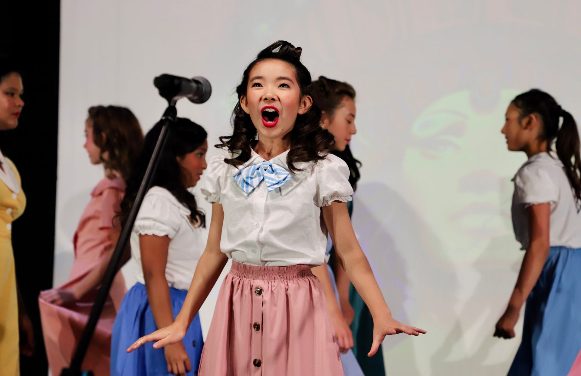 Girl singing on stage with other performers in the background for a theater performance.