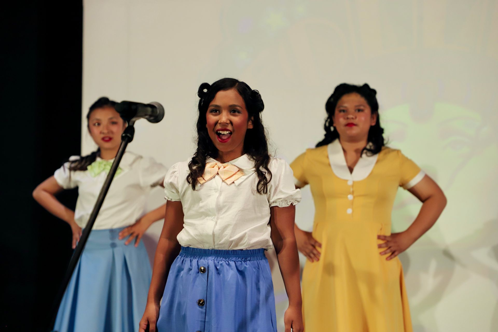 Three women singing onstage, wearing white tops with blue, white, and yellow skirts for a theater performance.