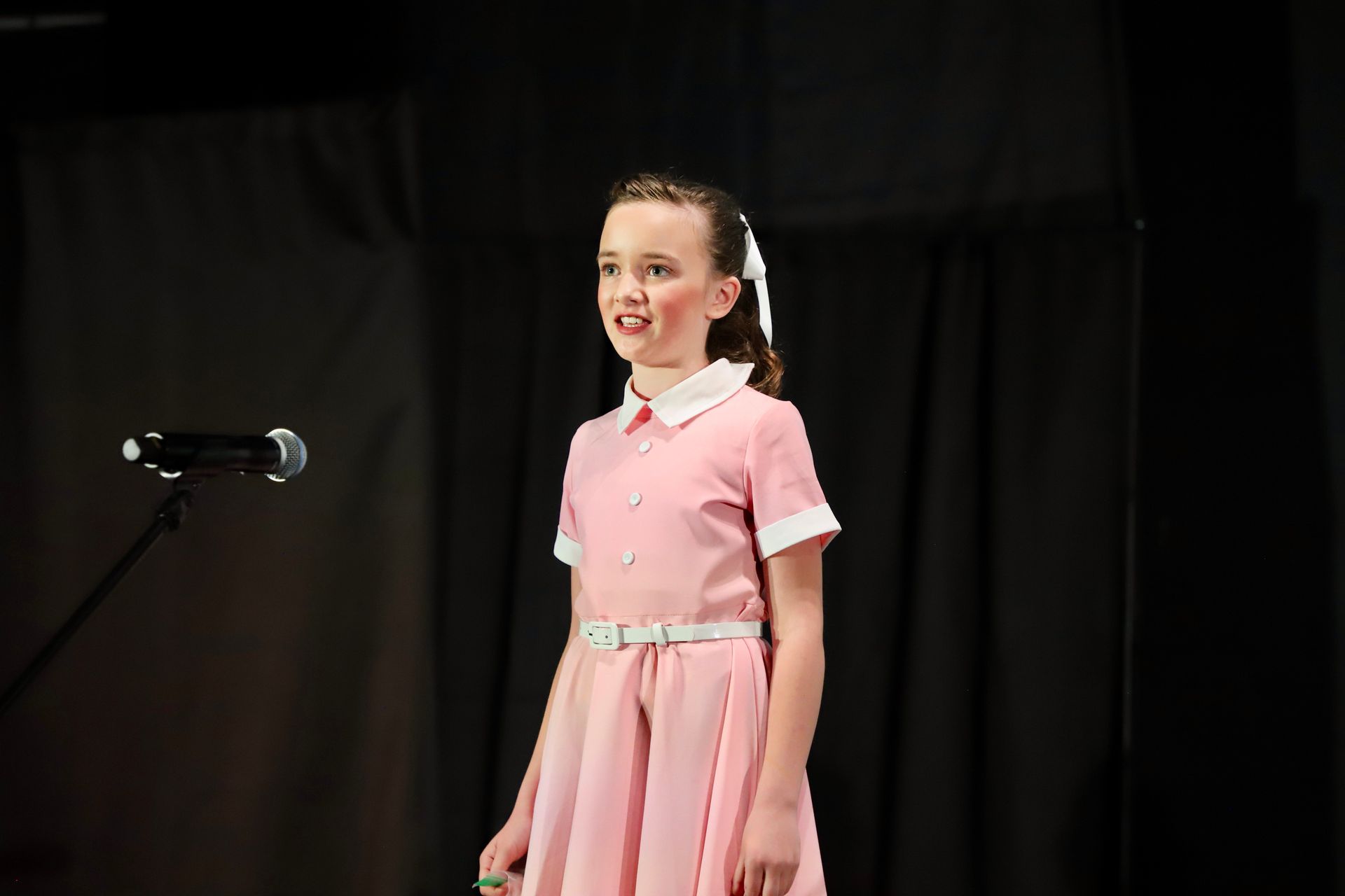 Young child in a pink dress speaking on stage beside a microphone, with a dark curtain backdrop for a theater performance.
