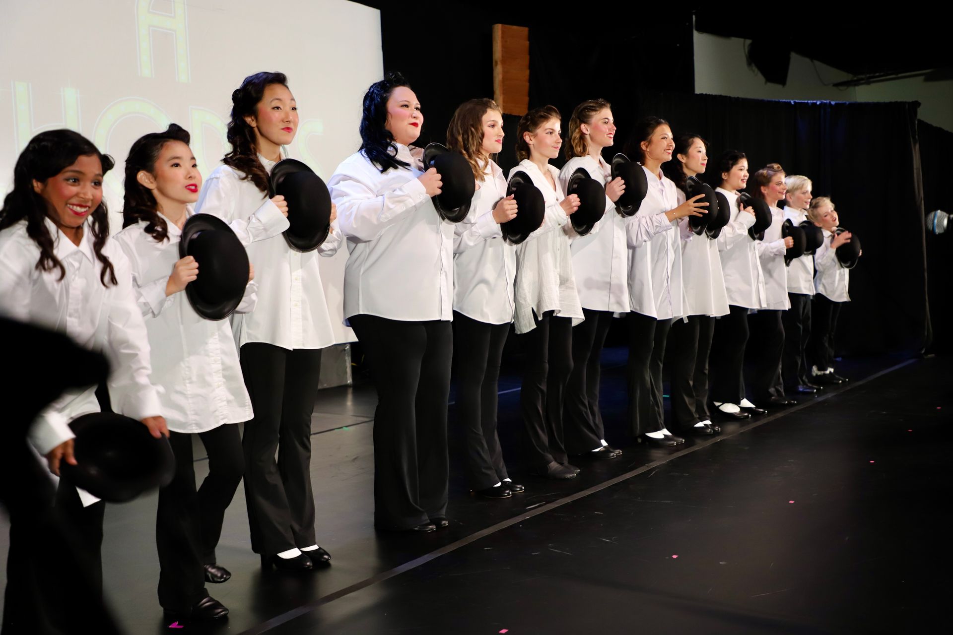 Row of performers in white tops and black pants standing onstage, holding black props  for a theater performance.