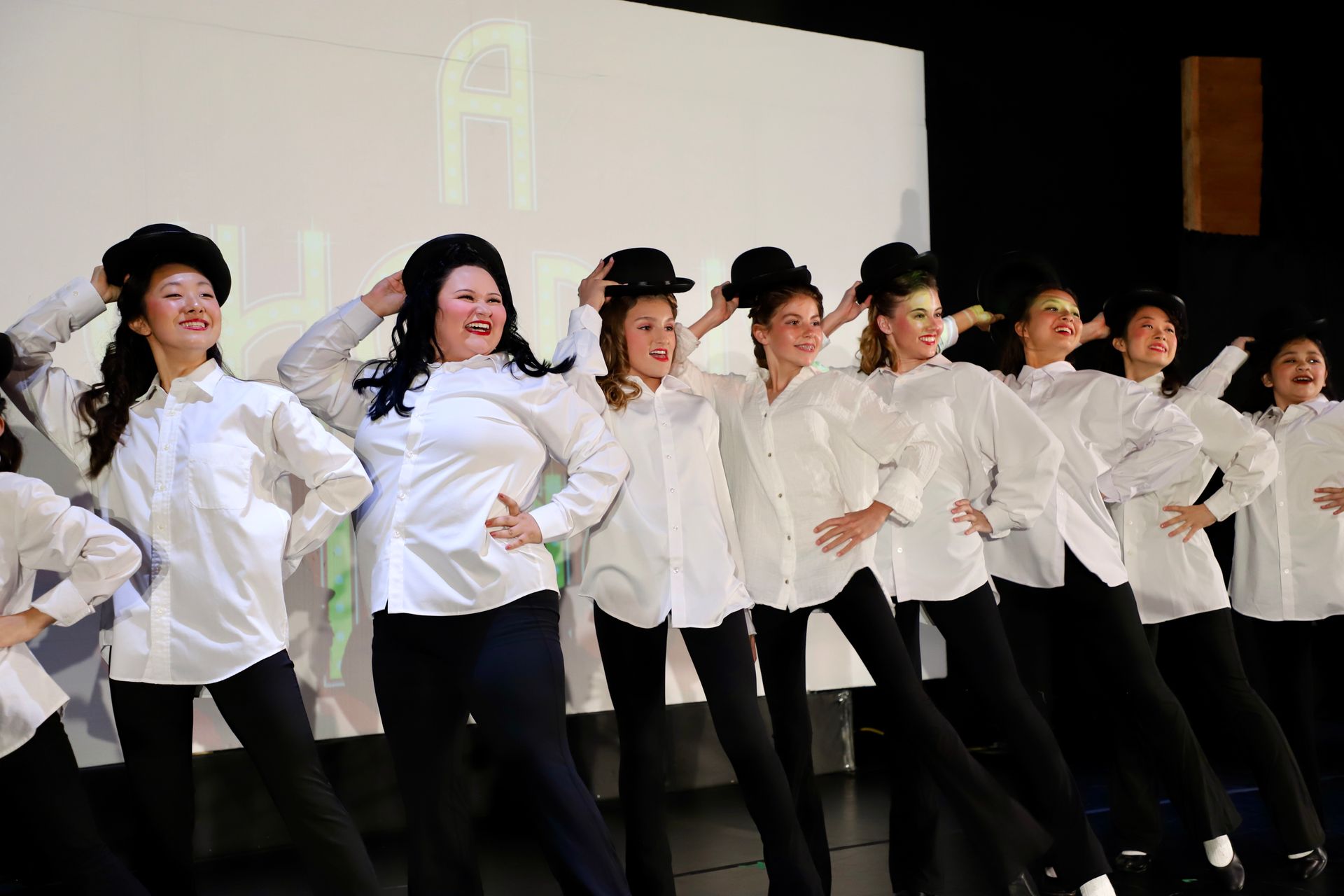 Performers in white shirts and black pants dancing in a line onstage under bright lights for a theater performance.