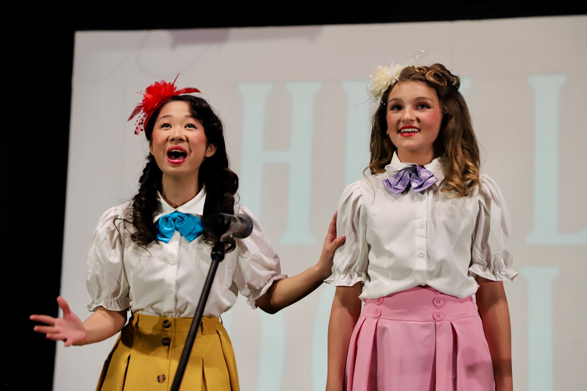 Two women singing onstage in white blouses and colorful skirts, smiling at microphones for a theater performance.