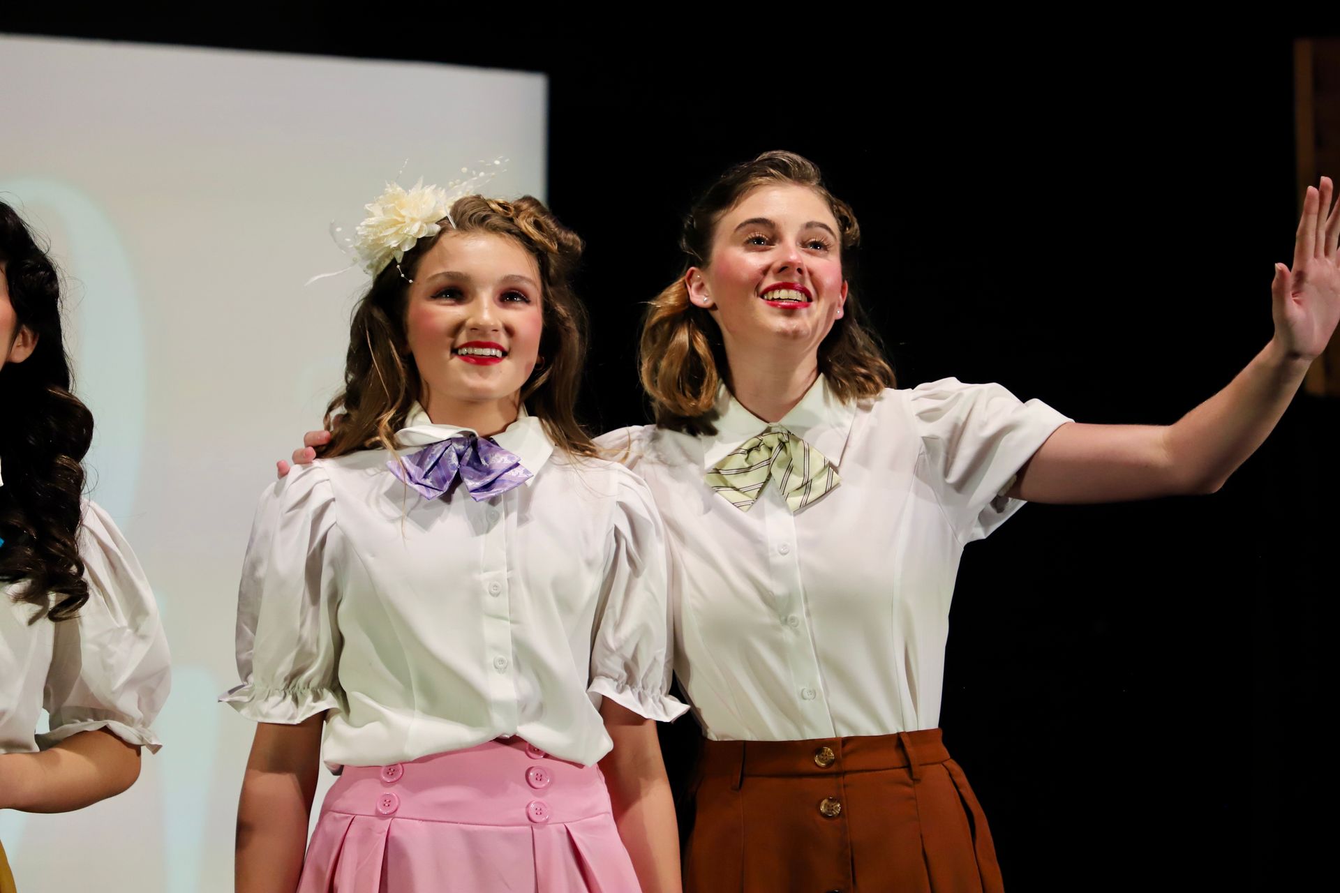 Two smiling performers in white blouses and colorful skirts waving on stage for a theater performance.