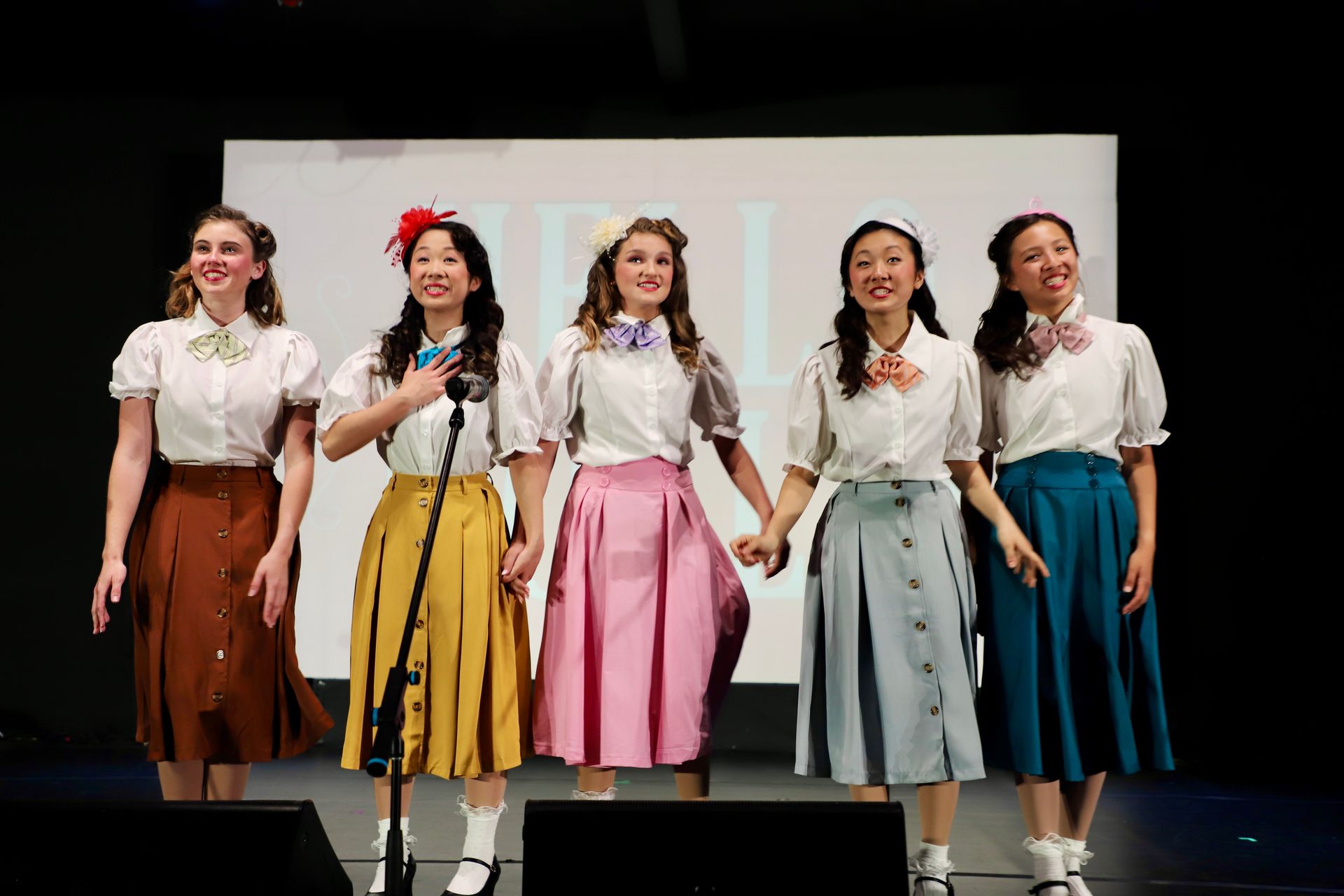 Five performers in colorful skirts sing onstage in front of microphones for a theater performance.