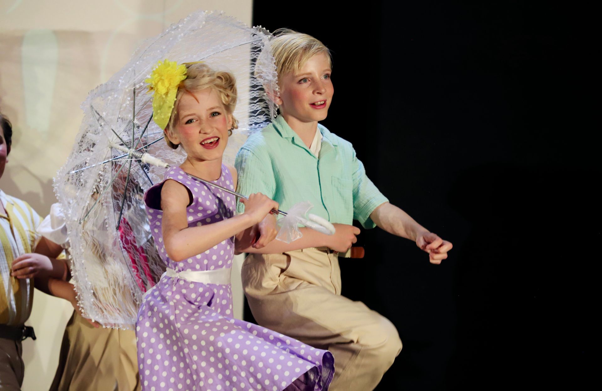 Two children in pastel costumes smile and dance onstage, one holding a white parasol for a theater performance.