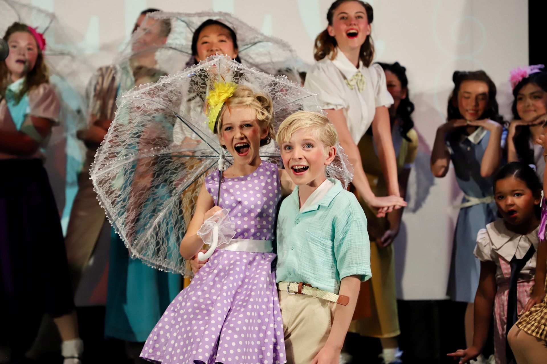 Children on stage in colorful costumes, smiling and posing with a lace parasol for a theater performance.