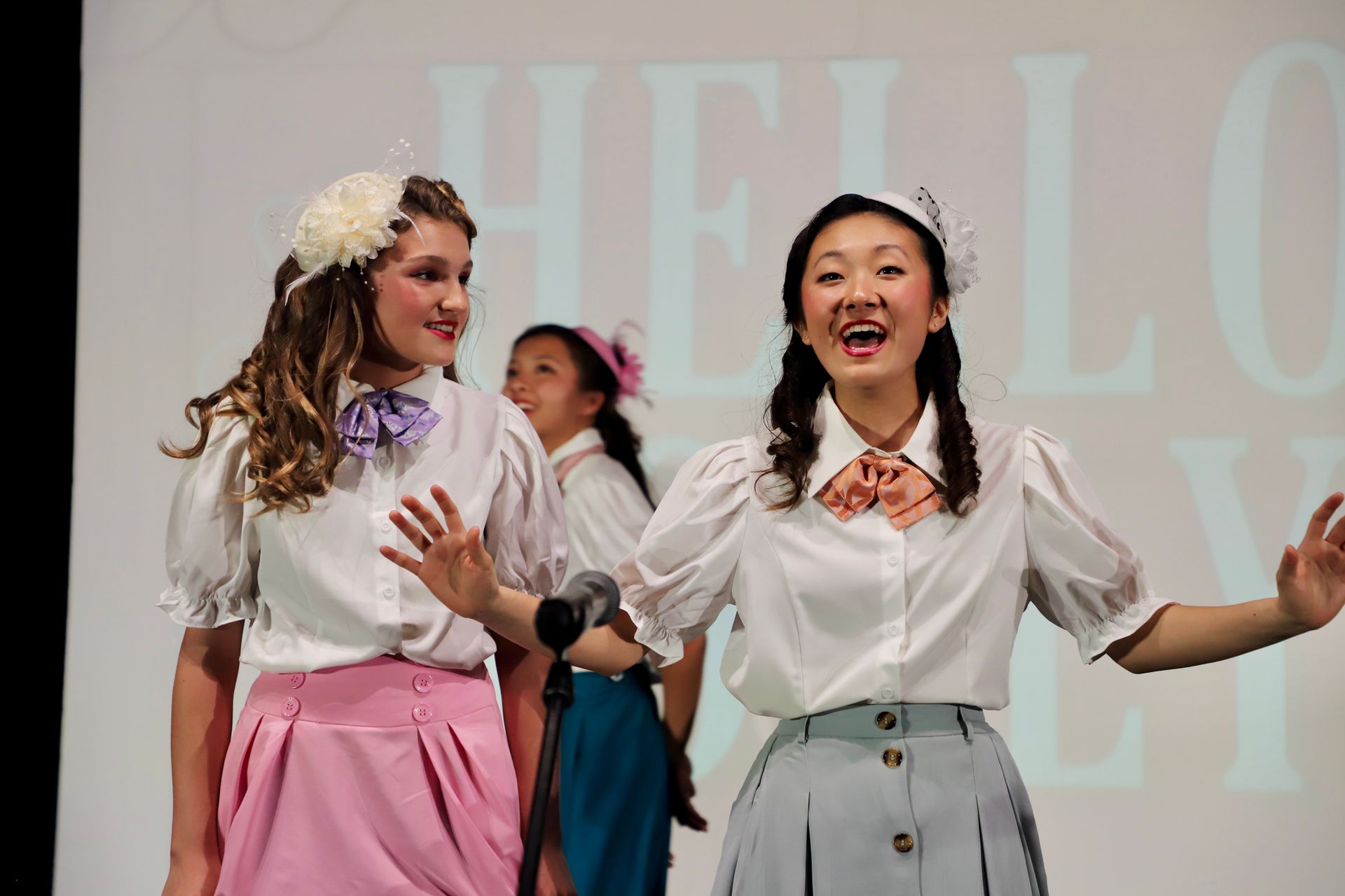 Two performers in white blouses and pink skirts sing onstage in front of a “HELLO” backdrop for a theater performance.