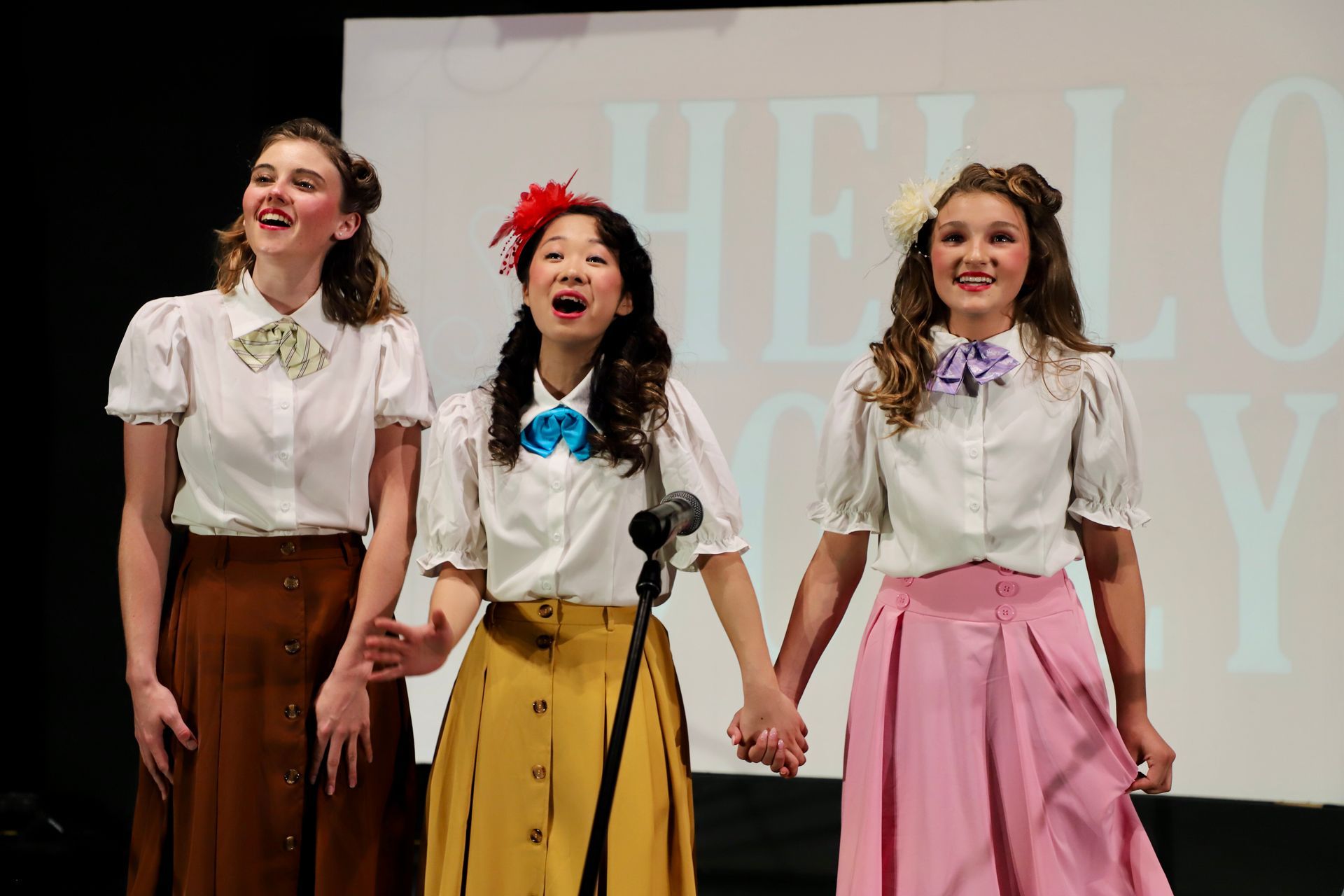 Three women on stage singing into microphones in front of a large “HELLO” backdrop for a theater performance.