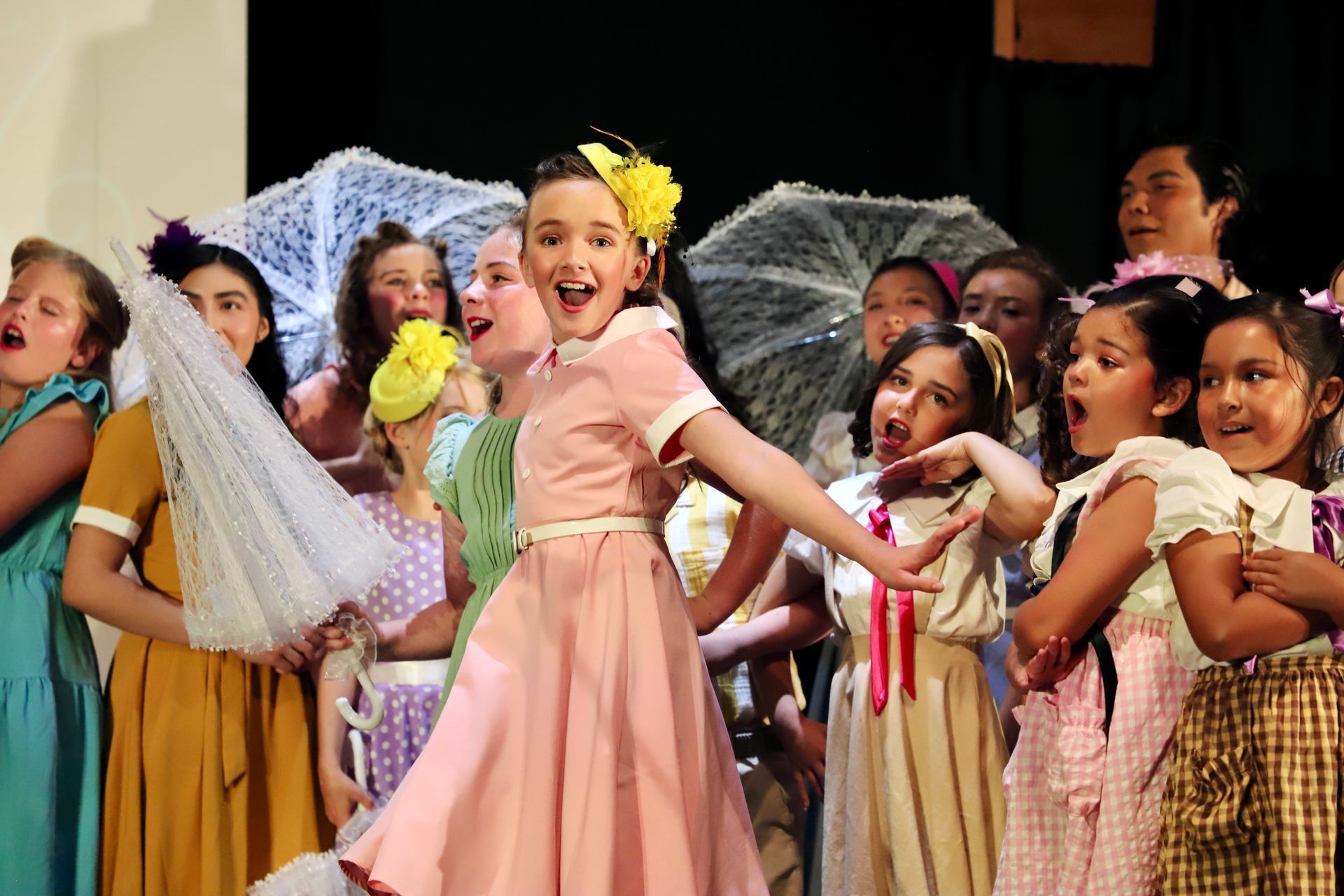 Children in colorful dresses perform on stage, smiling and holding lace props for a theater performance.
