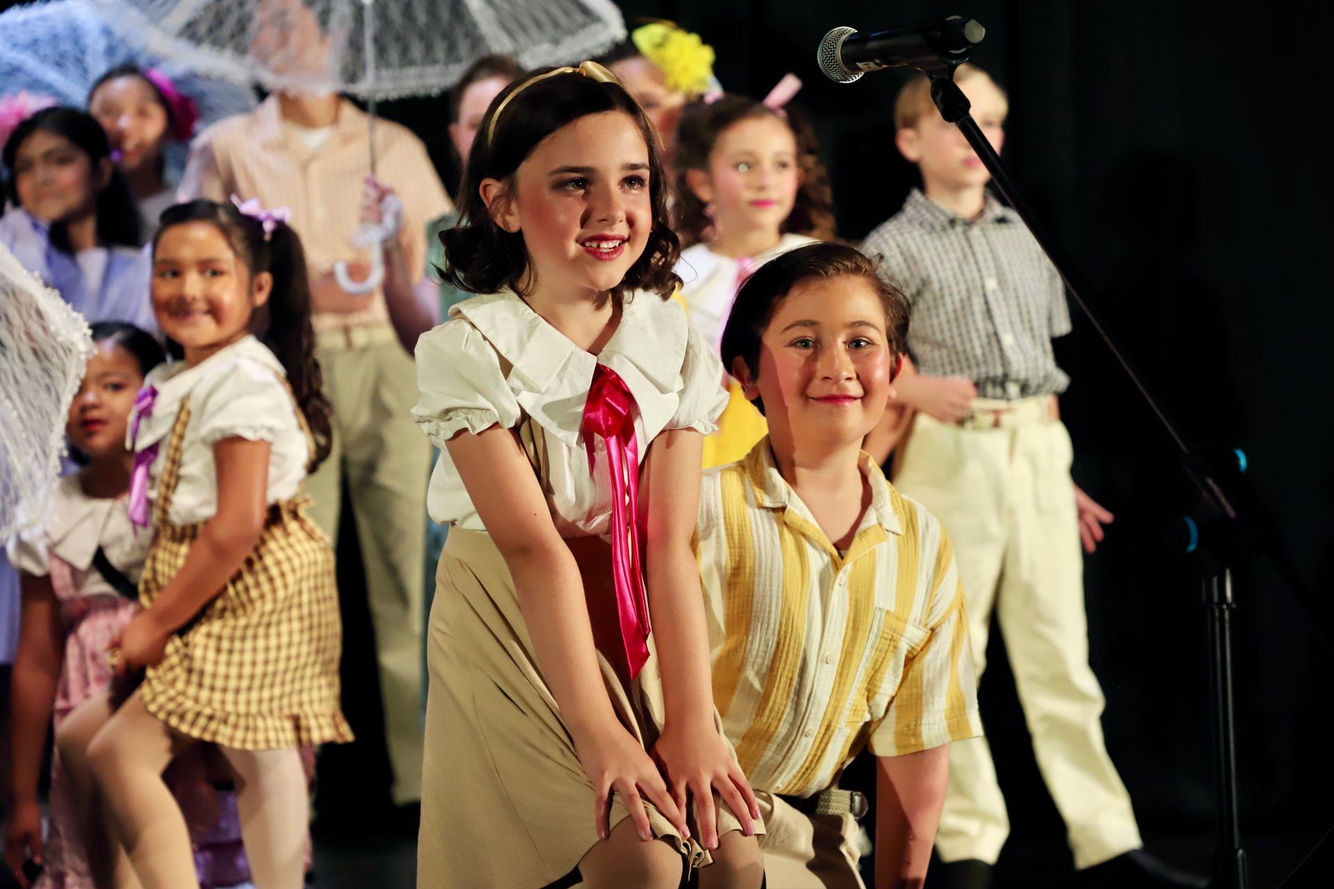 Children performing a dance onstage, smiling in colorful costumes with a dark backdrop for a theater performance.