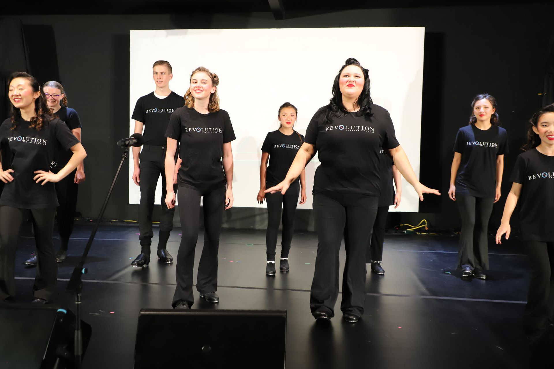Group of performers in black shirts standing on a stage in front of a screen for a theater performance.