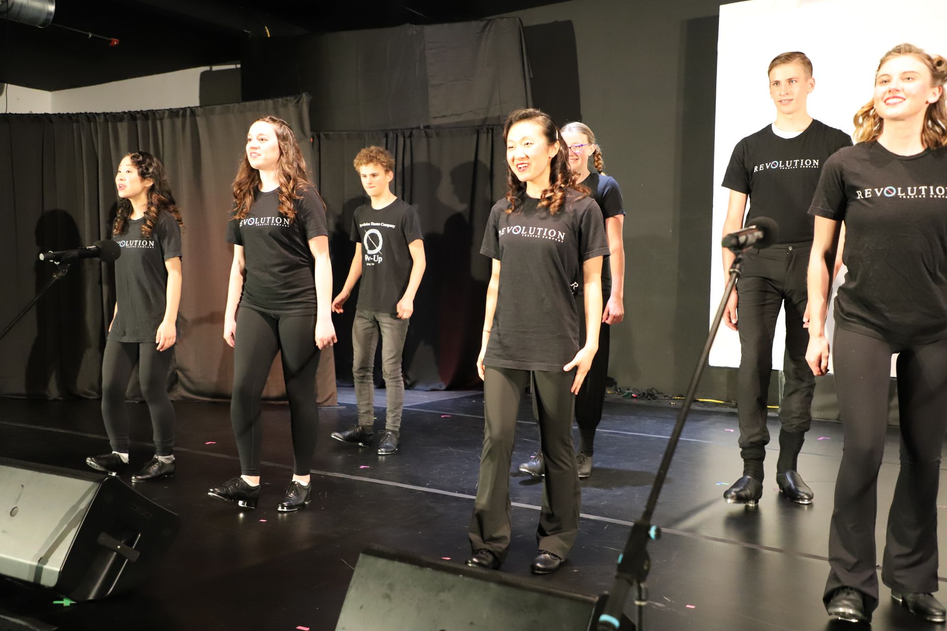 Group of performers in black outfits standing on a stage with microphones and speakers for a theater performance.