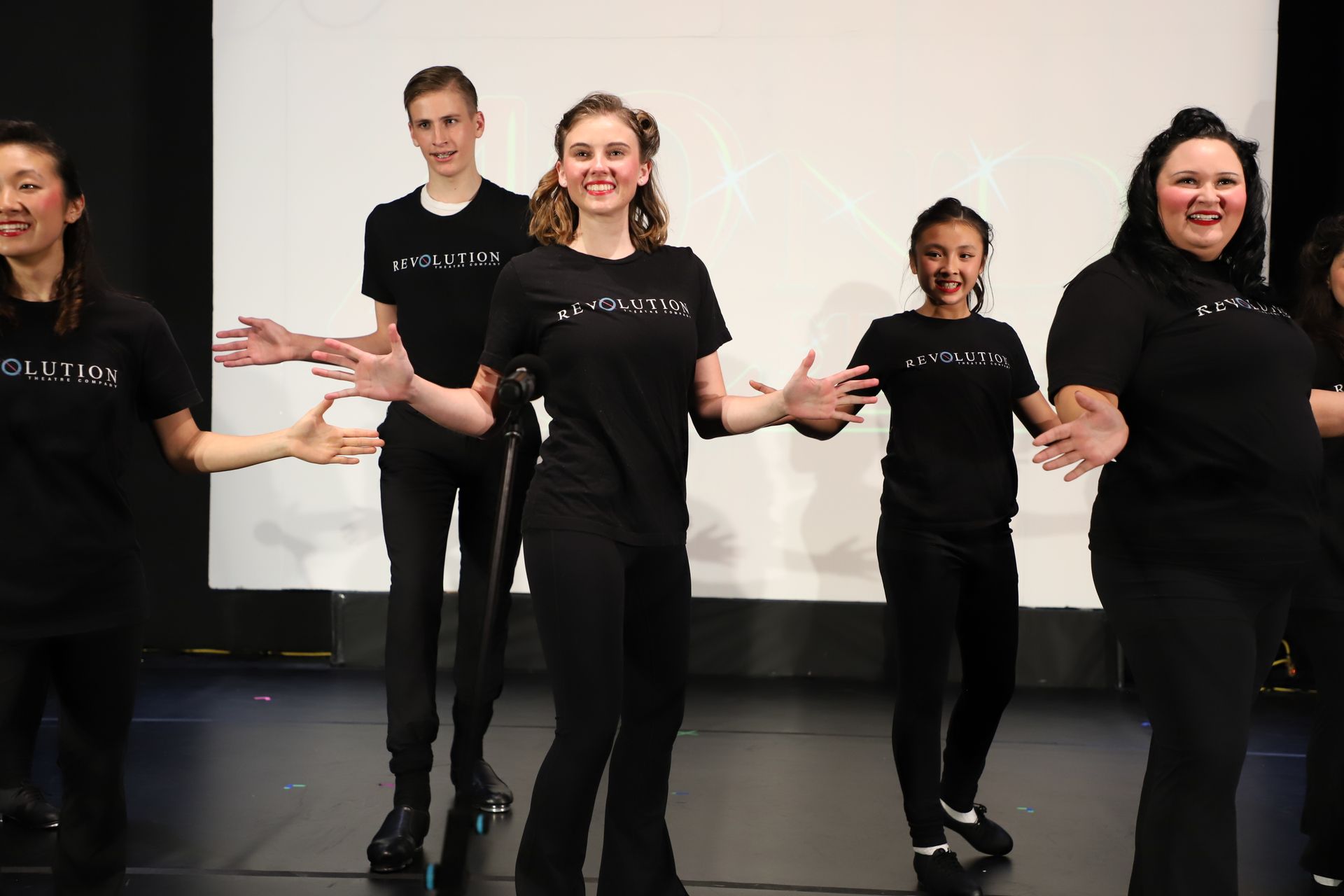 Five people in black shirts standing on a stage, smiling and gesturing with open hands for a theater performance.