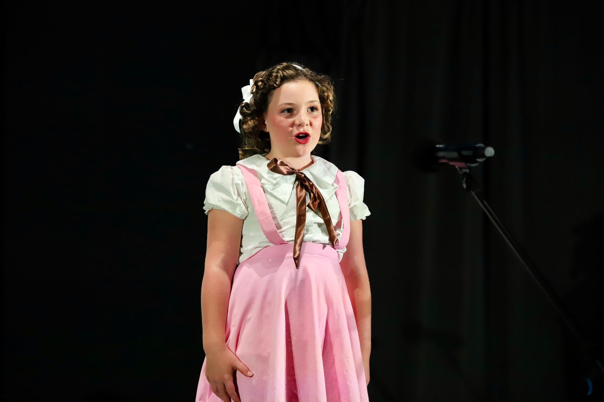 Young performer in a pink dress singing onstage into a microphone against a dark background for a theater performance.