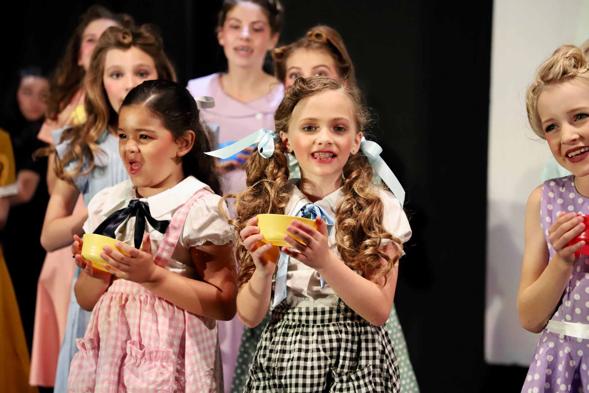 Children in sparkly dresses holding small trophies on a stage during a performance for a theater performance.