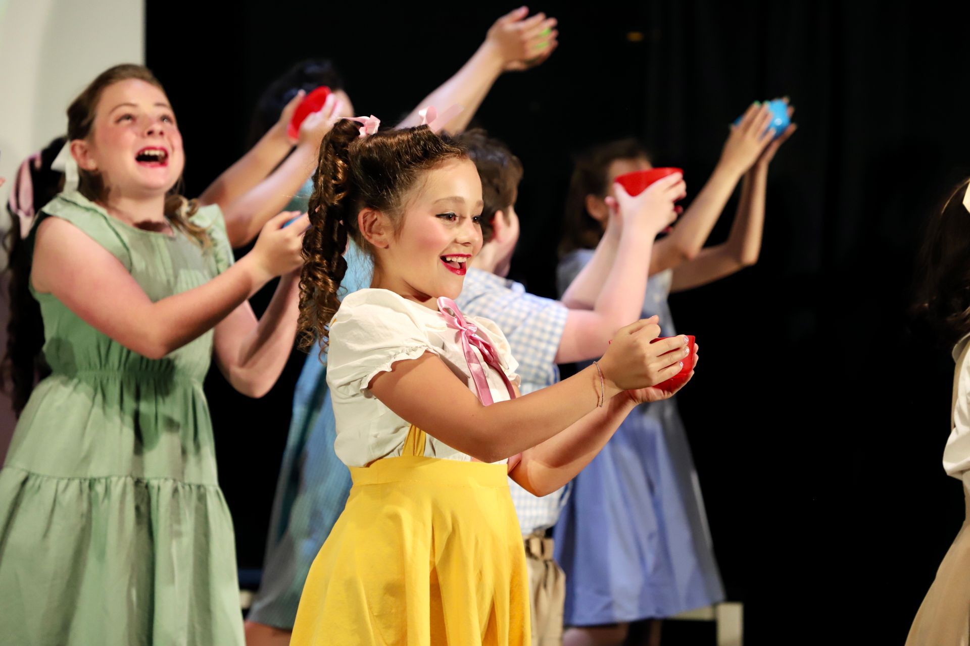 Children performing a colorful dance on stage, smiling and clapping with scarves raised for a theater performance.