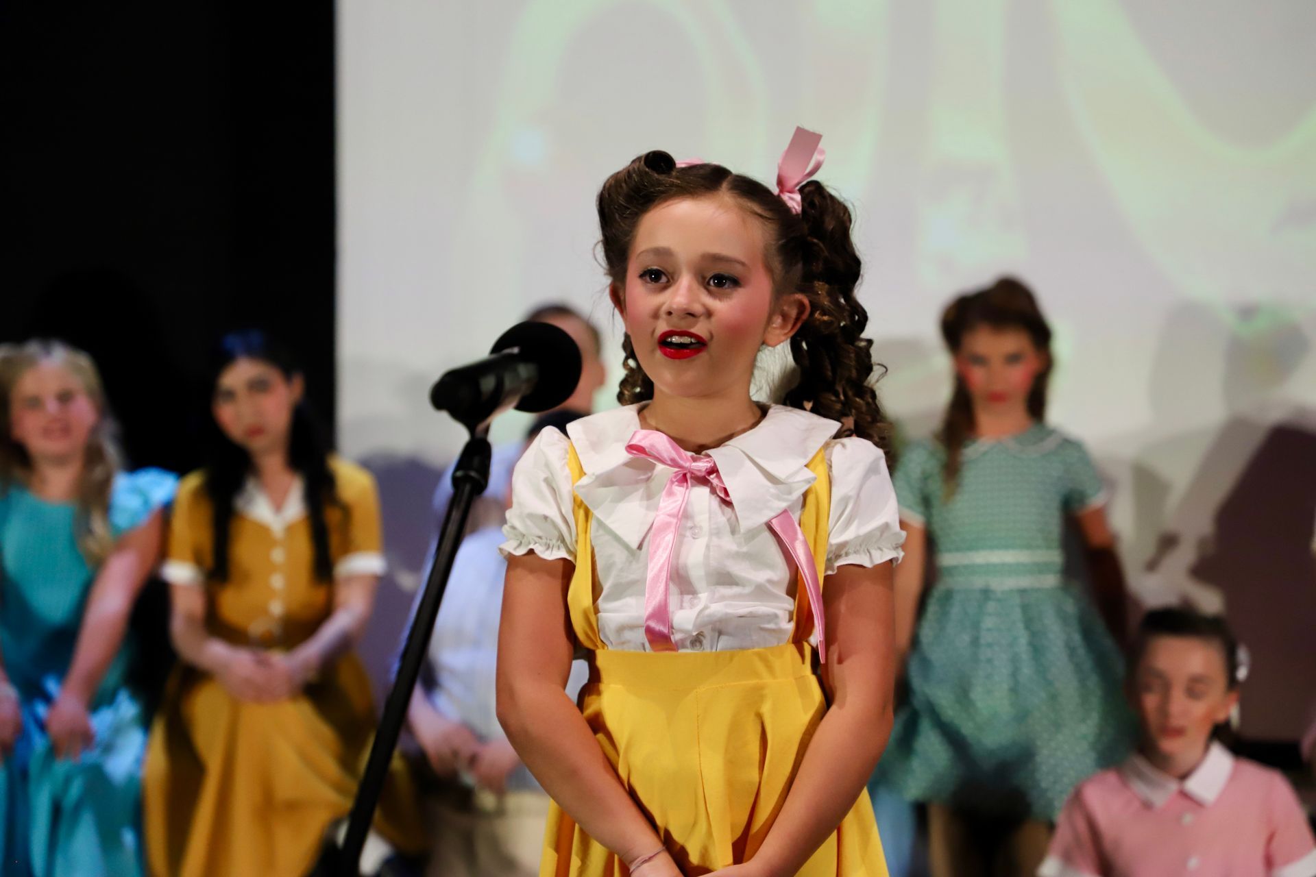 Child in yellow skirt singing into a microphone onstage, with other performers in the background for a theater performance.