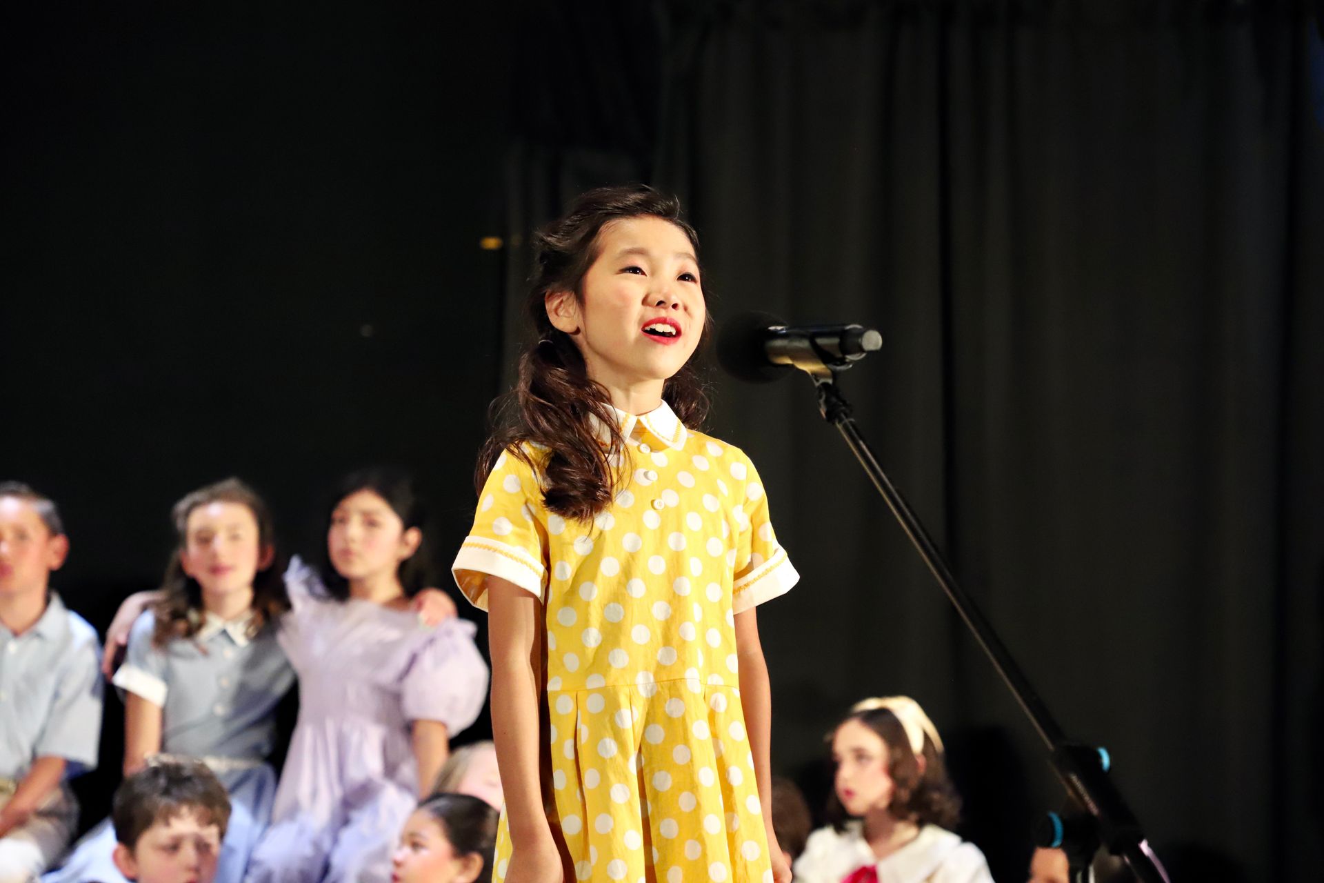 Child in a yellow dress singing into a microphone on stage with a choir behind her for a theater performance.