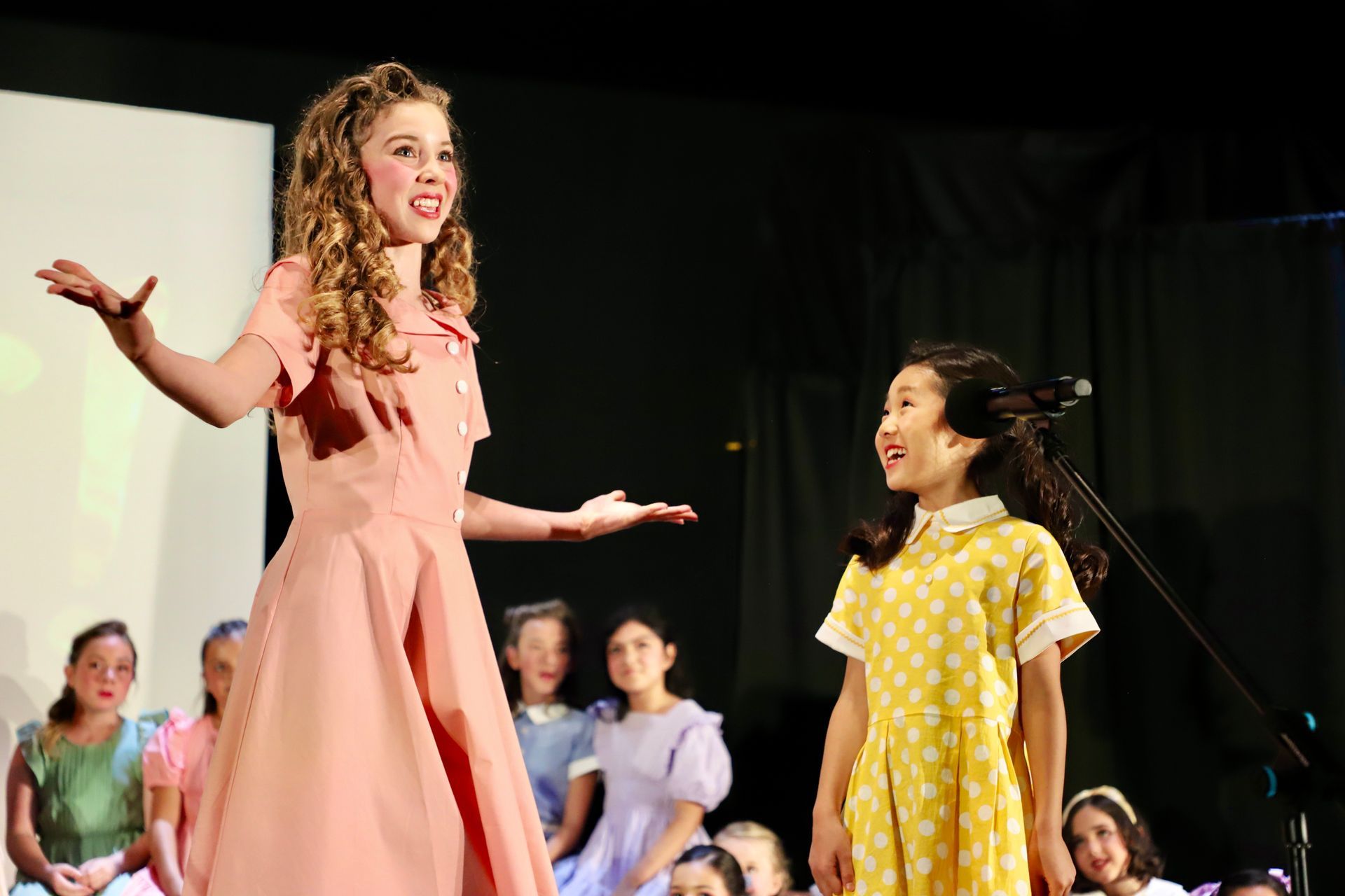 Two performers onstage in colorful dresses, gesturing and smiling during a live performance for a theater performance.