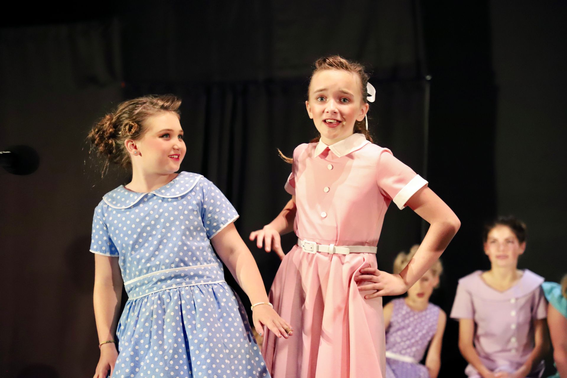 Two girls in pastel dresses pose onstage; one smiles with hands on hips for a theater performance.