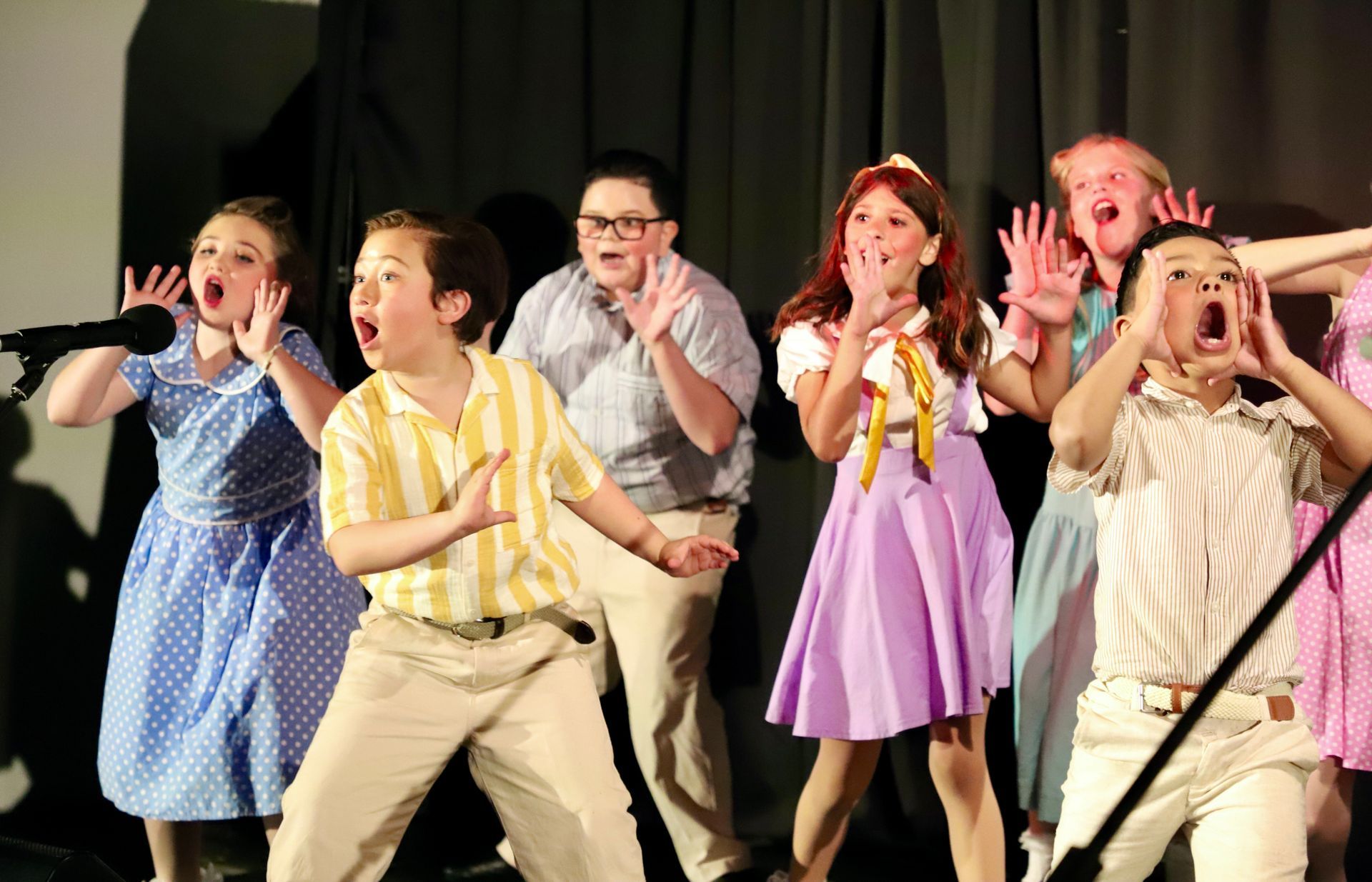 Performers on stage singing and dancing, holding microphones in costumes under bright lights for a theater performance.