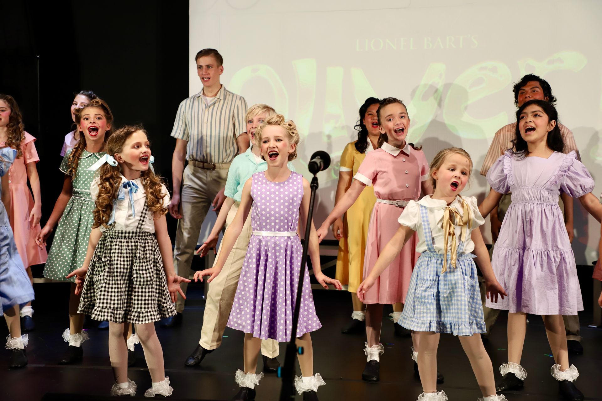 Children performing on stage in colorful costumes, standing in formation with arms outstretched for a theater performance.