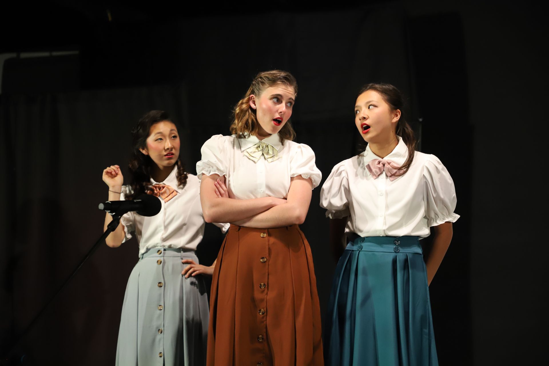 Three performers onstage in white blouses and long skirts, posing and singing under stage lights for a theater performance.