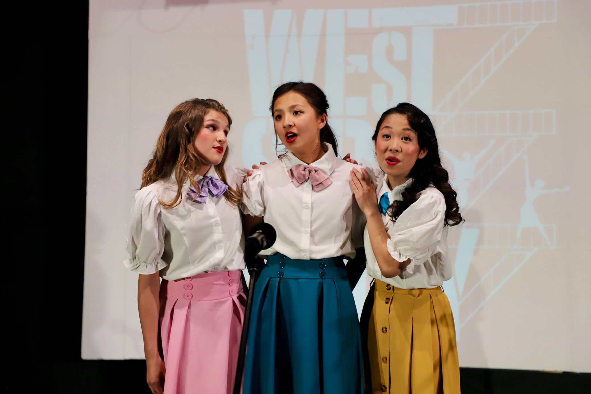 Three women singing onstage in white blouses and colorful skirts, standing before a lit screen for a theater performance.