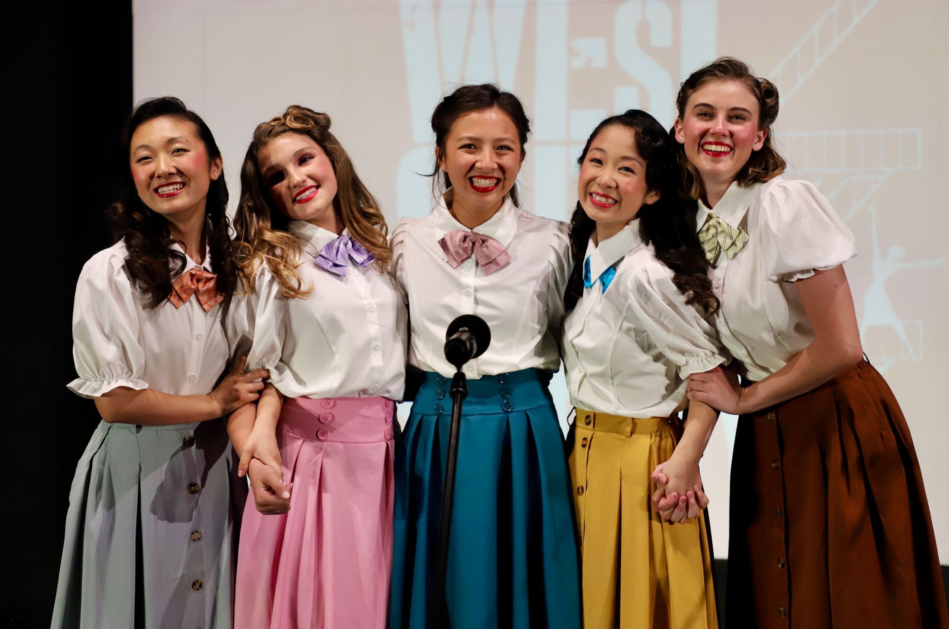 Five smiling performers in white blouses and colorful skirts stand arm in arm on stage for a theater performance.