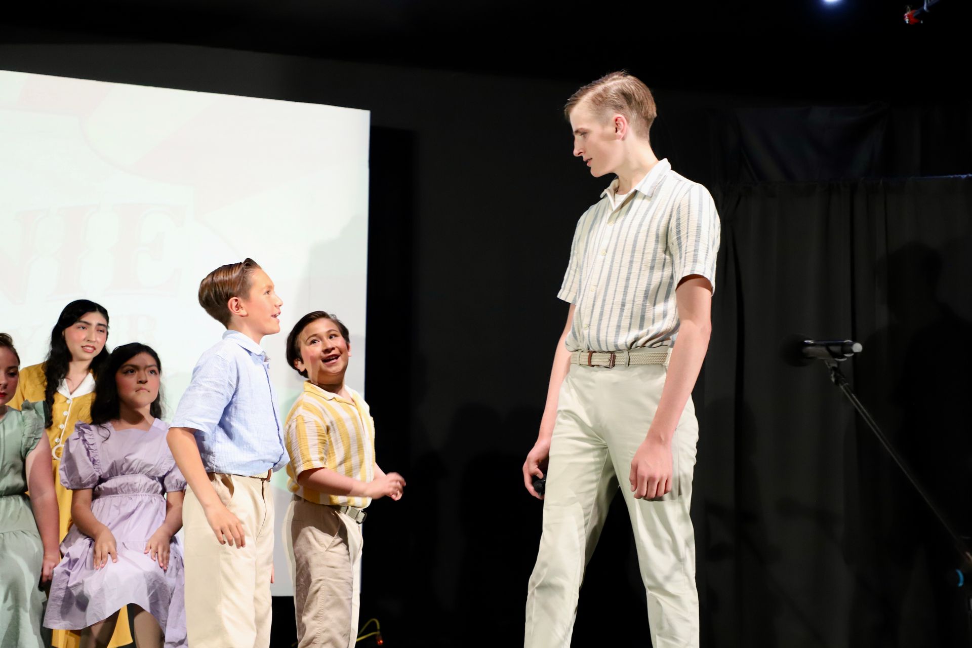 Children on stage facing a tall man in a plaid shirt under bright theater lights for a theater performance.