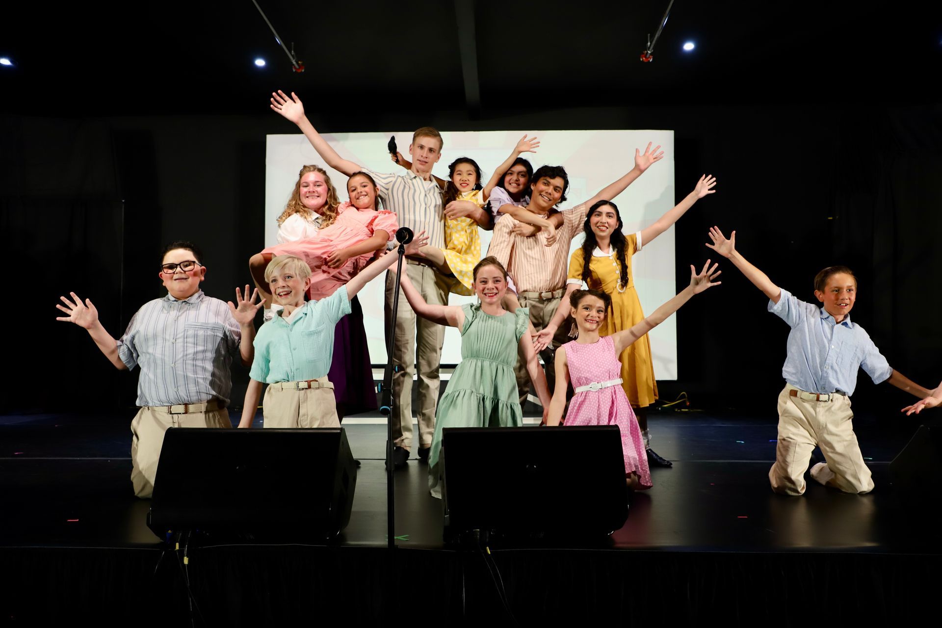 A group of performers pose on a stage with raised arms, under spotlights and in colorful outfits for a theater performance.