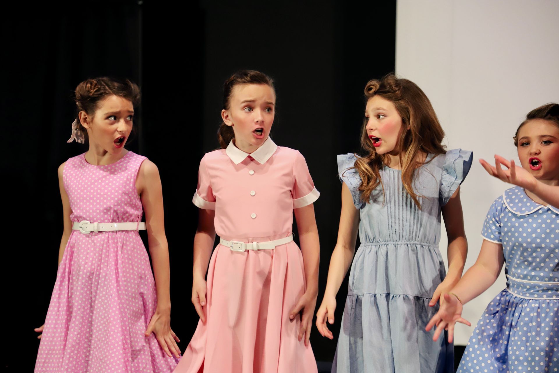 Four children in pastel dresses onstage, singing with surprised expressions for a theater performance.