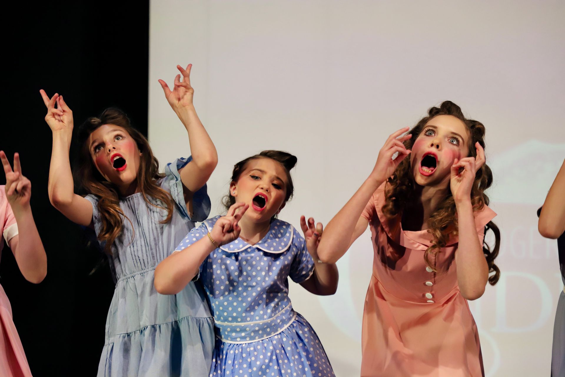 Children onstage in colorful dresses, singing and raising their hands during a performance for a theater performance.