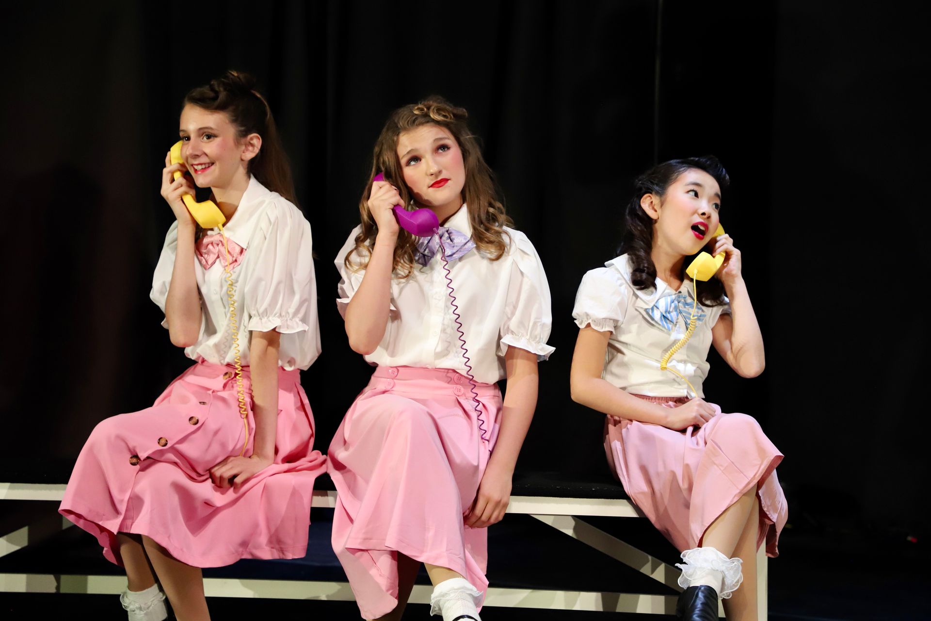 Three performers in pink skirts and white tops sit onstage holding yellow phones for a theater performance.