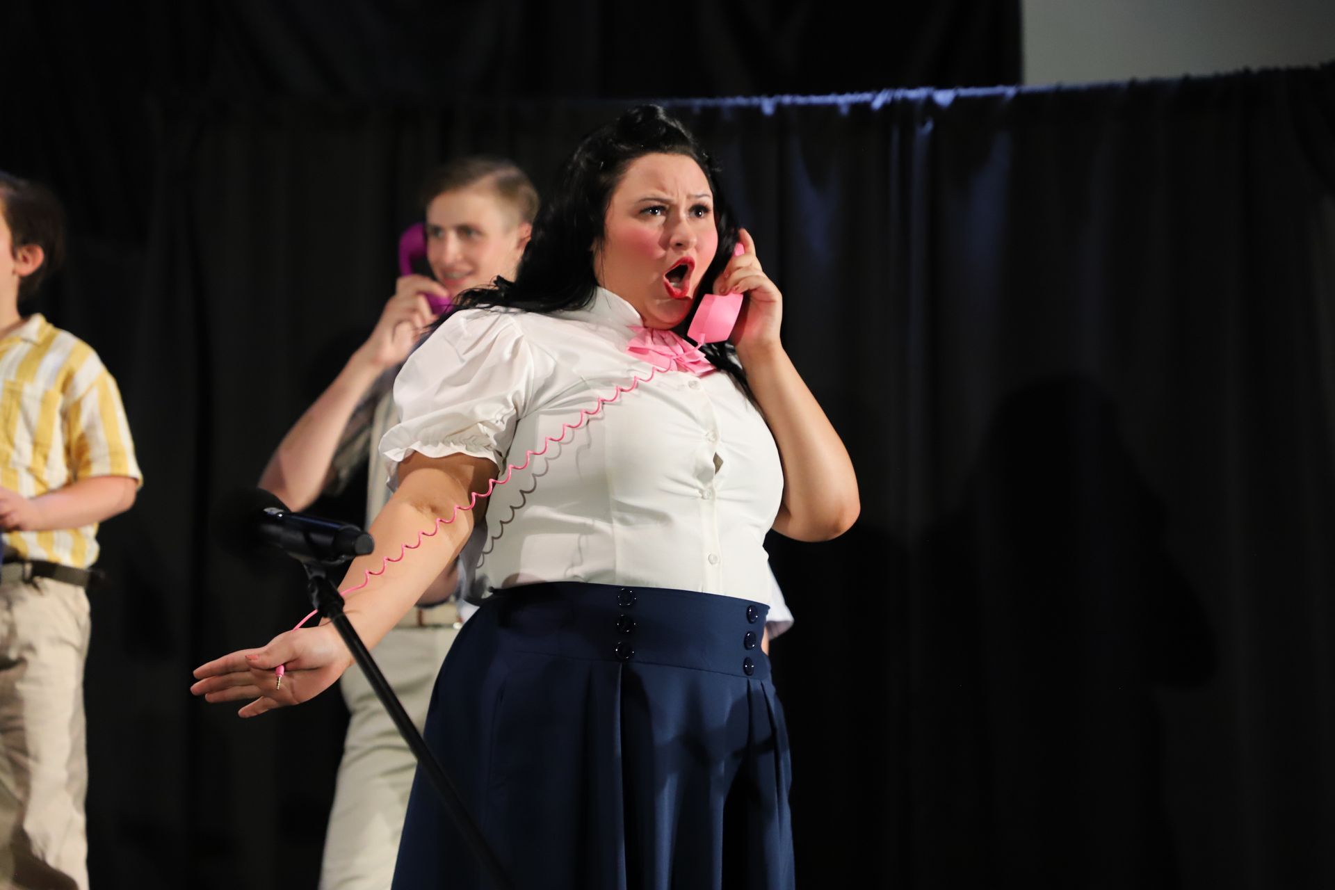Woman in white top and navy skirt speaking into a pink phone onstage  for a theater performance.