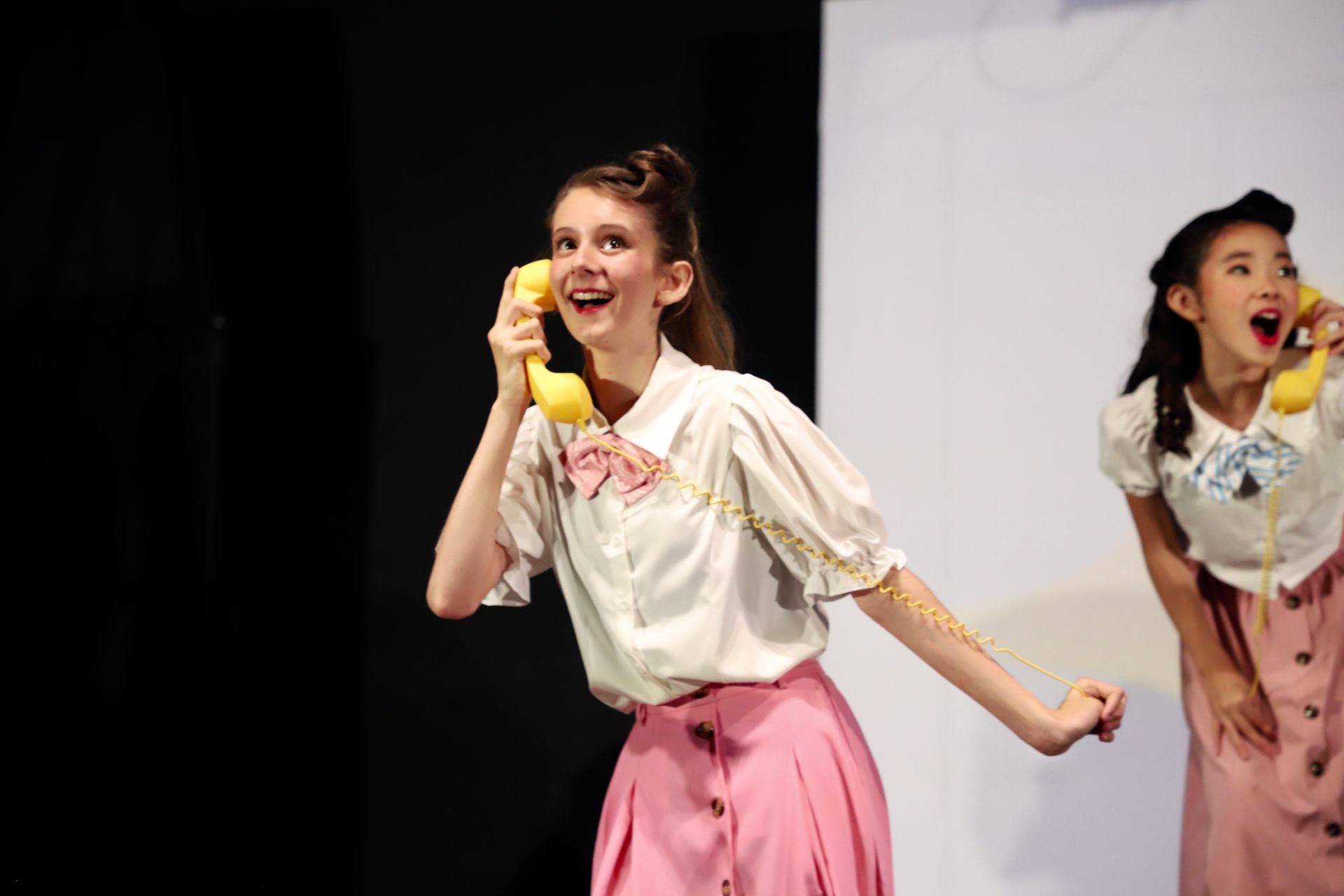 Two performers in white blouses and pink skirts sing onstage, one holding a yellow prop banana for a theater performance.