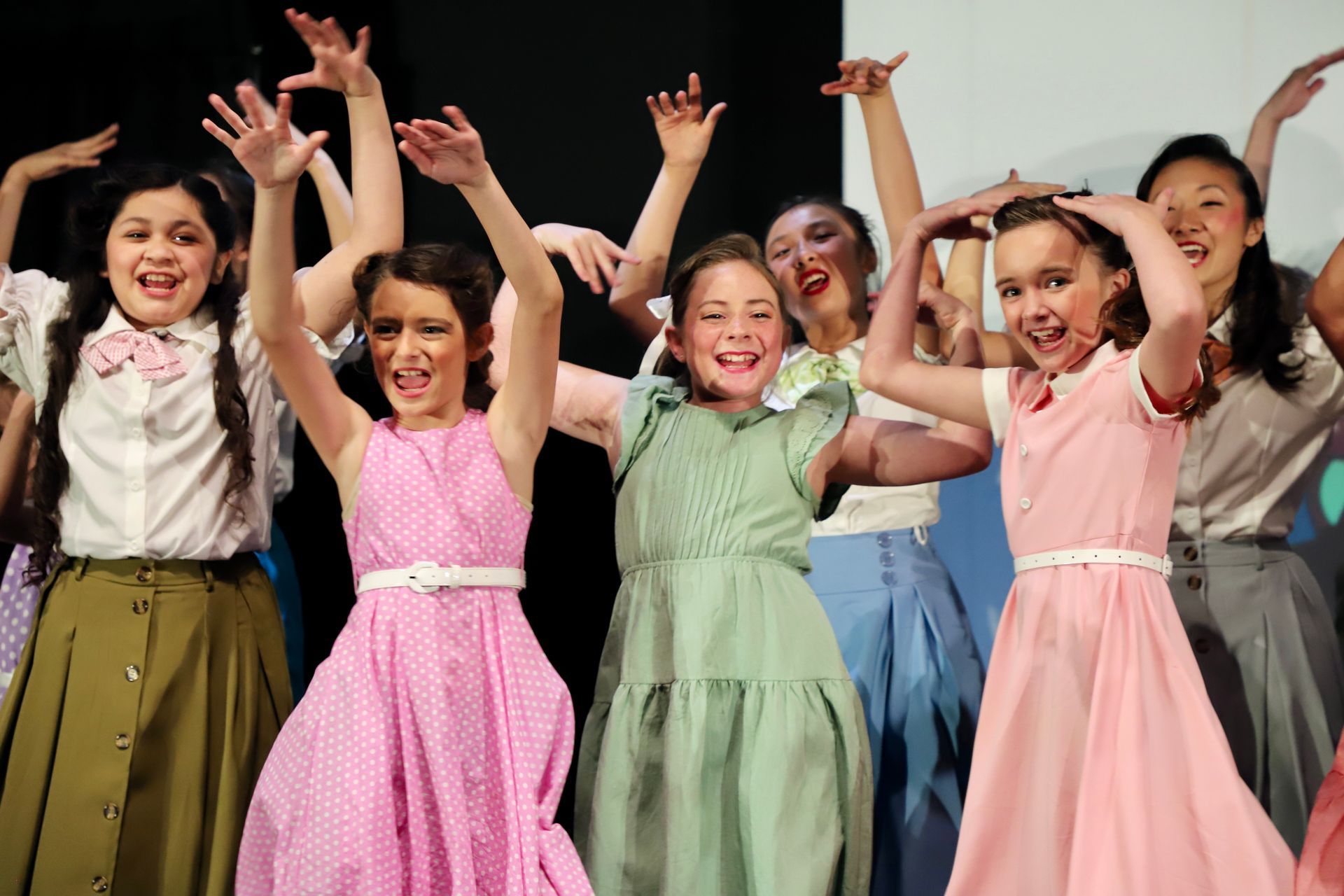 Children dancing on stage in colorful dresses with arms raised, smiling under stage lights for a theater performance.