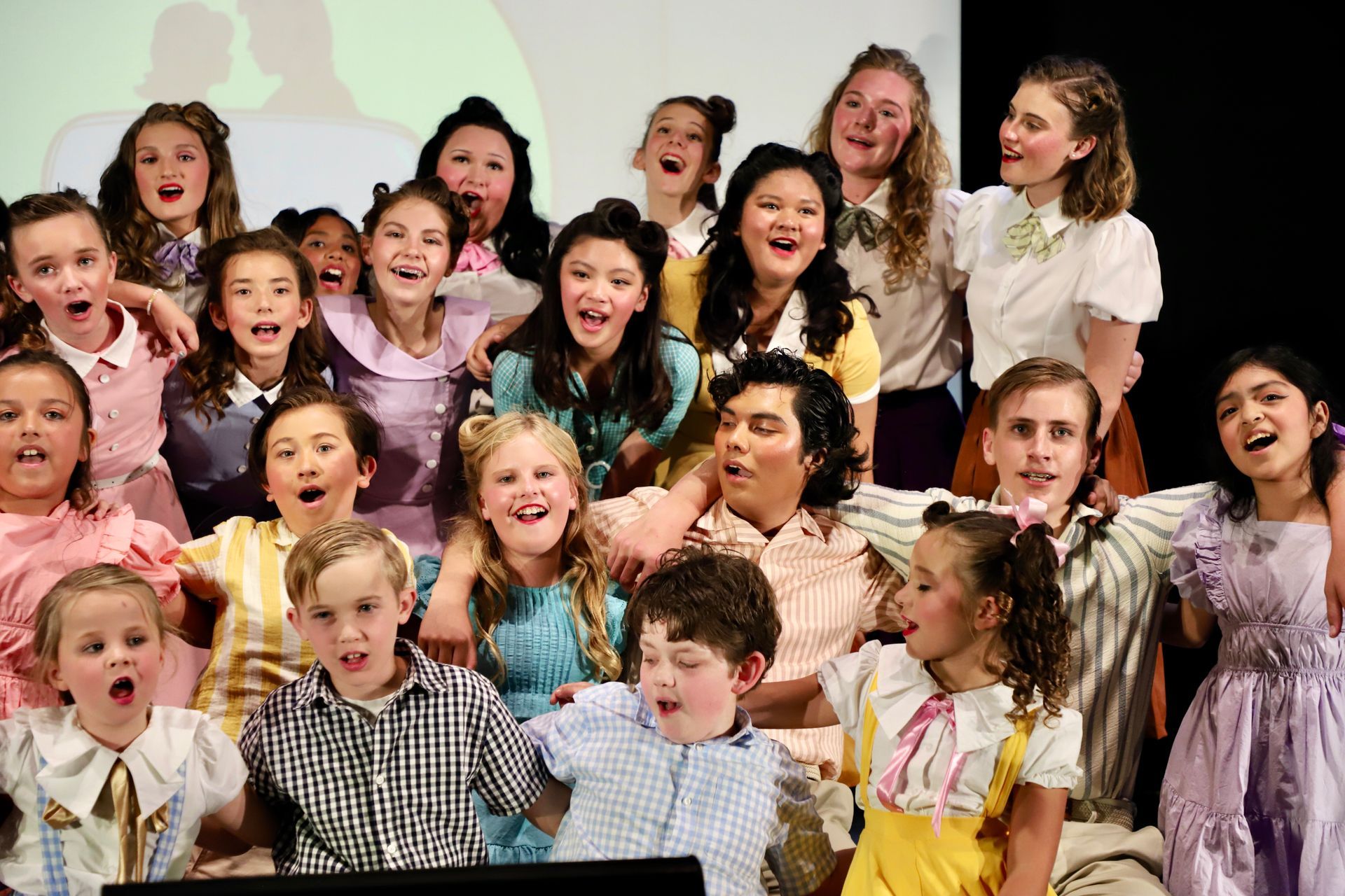 Children singing on stage, smiling and cheering in colorful clothing for a theater performance.