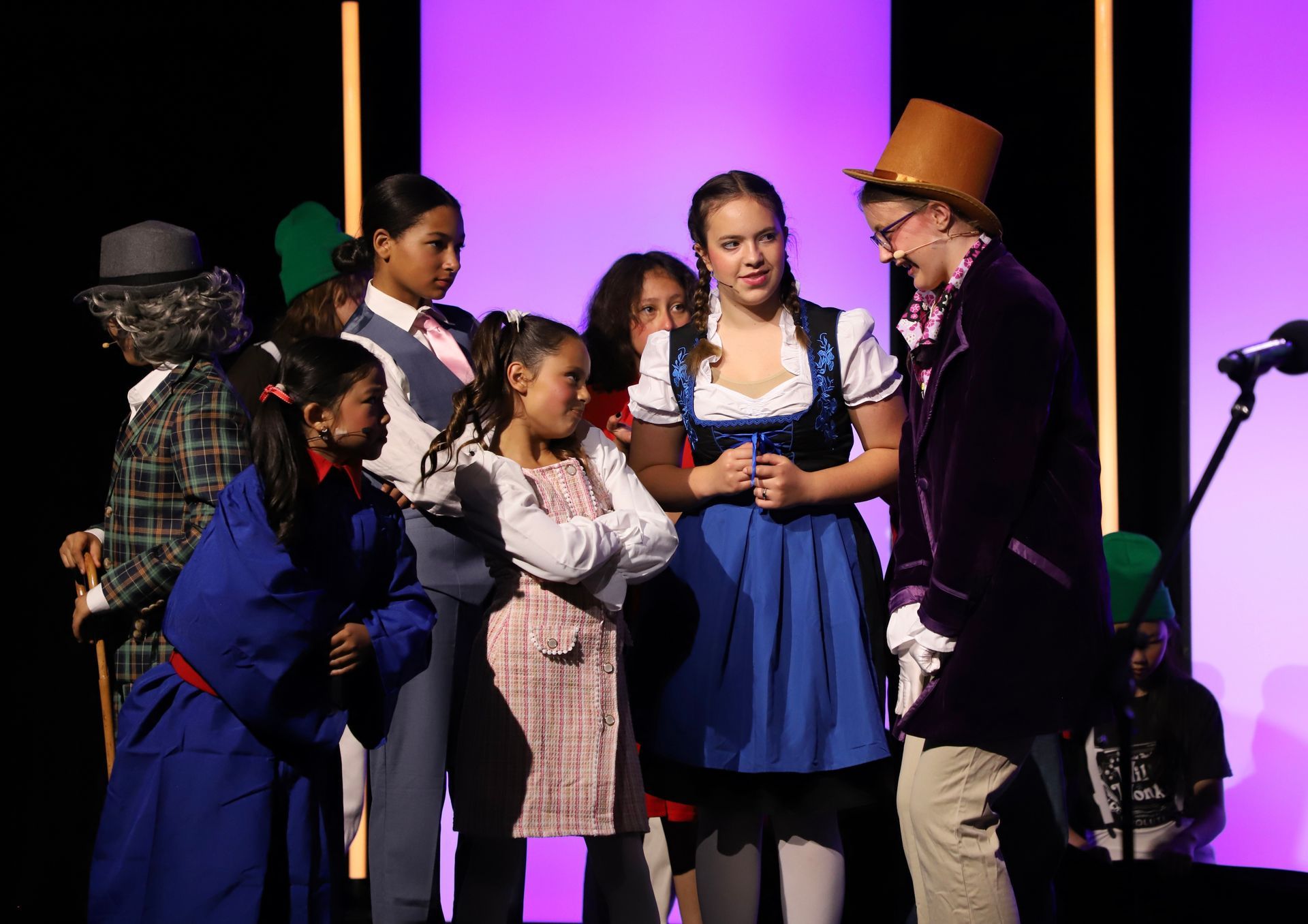 A group of performers in colorful costumes gather onstage around a girl in a blue dress for a theater performance.