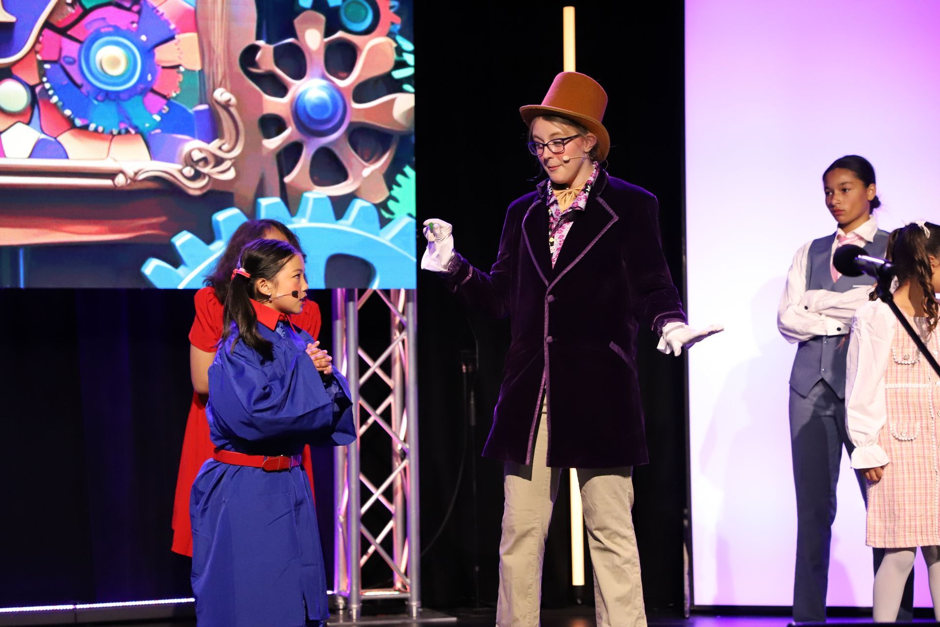 Three people on stage in costume, speaking beside a colorful projected backdrop for a theater performance.