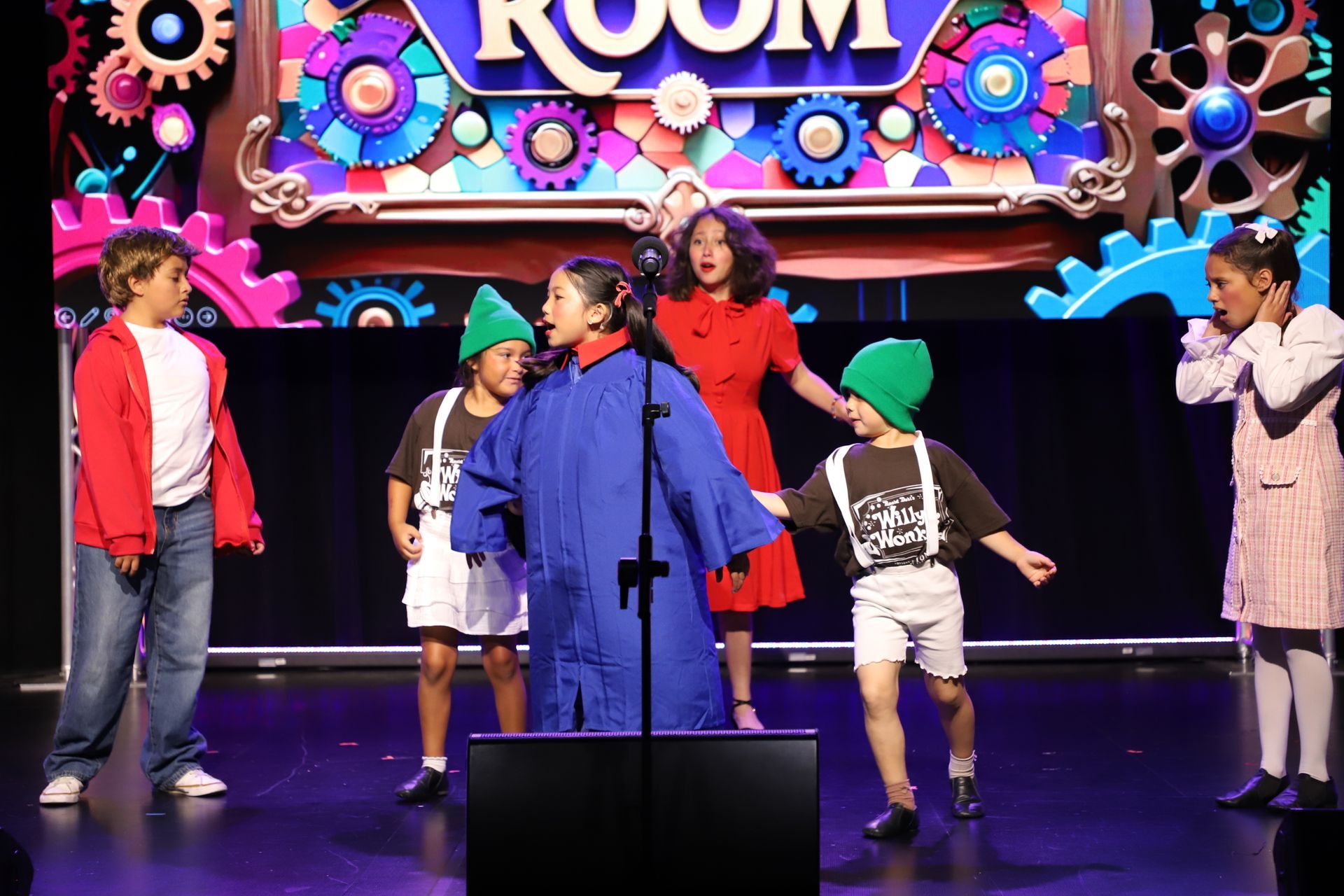 Performers onstage singing and dancing under colorful neon lights with a large “ROOM” backdrop for a theater performance.