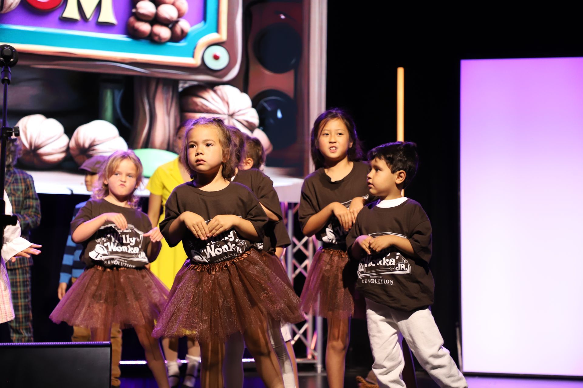 Four children perform a dance onstage in matching brown costumes under colorful stage lights for a theater performance.