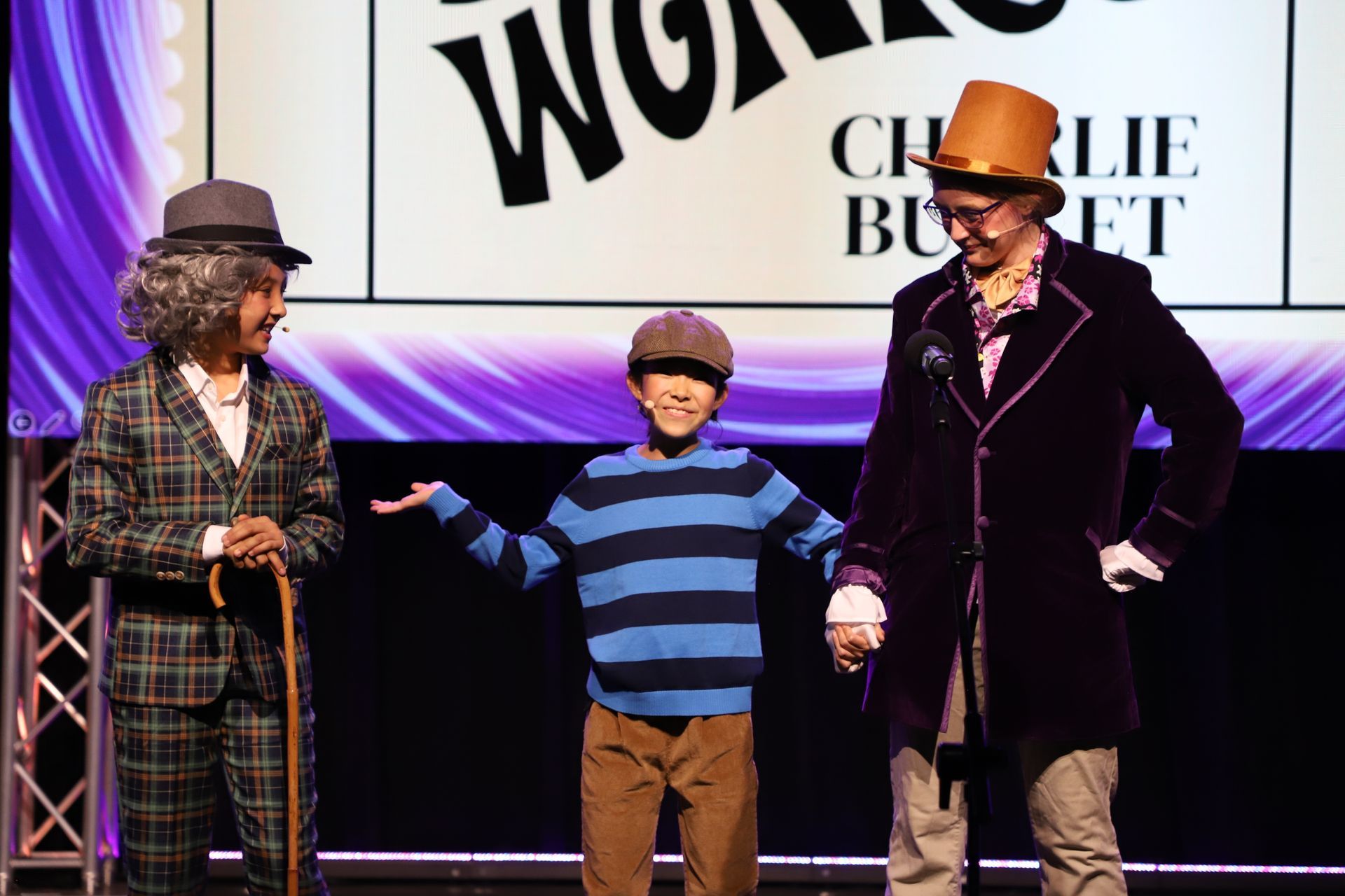 Three people on stage in costumes, standing before a Wonka-themed backdrop for a theater performance.