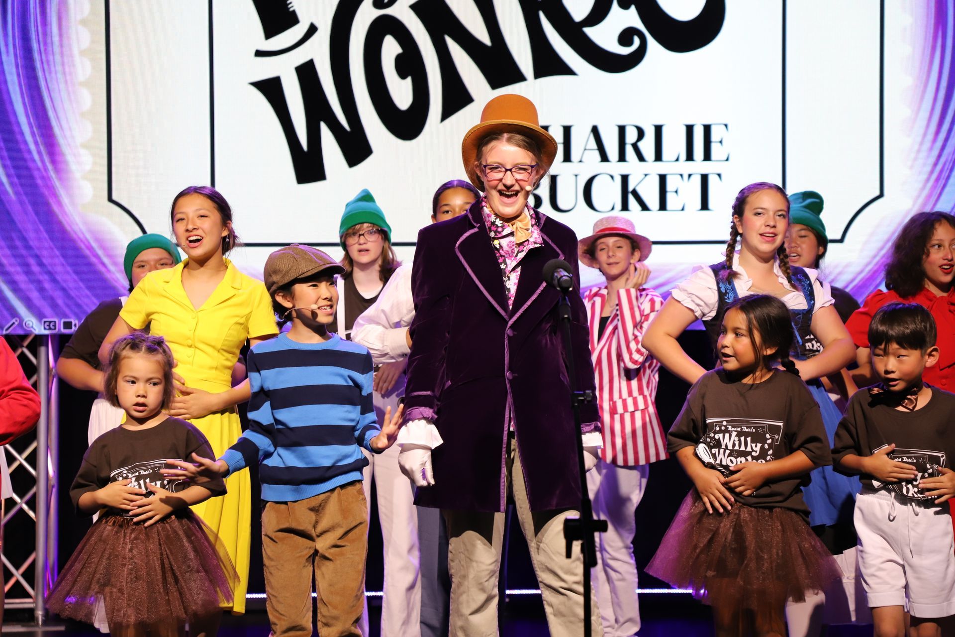 Children performing on stage at a Wonka-themed event, with a child in a purple velvet coat for a theater performance.