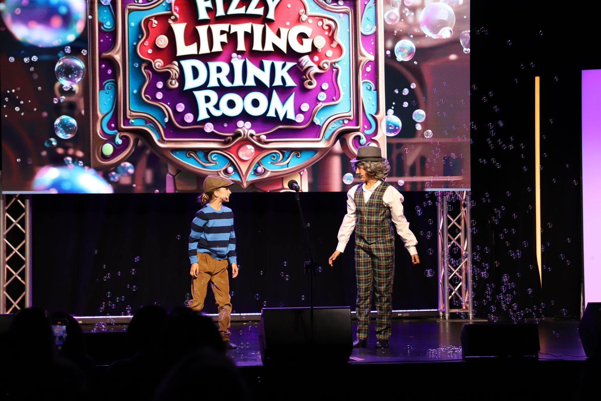 Two performers onstage beneath a colorful “Fizzy Lifting Drink Room” sign with sparkling lights for a theater performance.