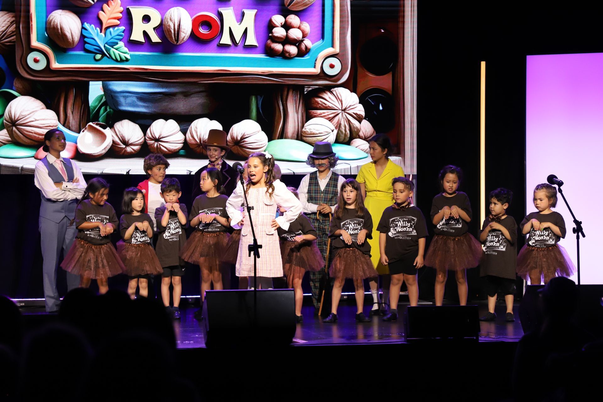 Children performing on stage in sparkly costumes with a colorful “ROM” sign behind them for a theater performance.