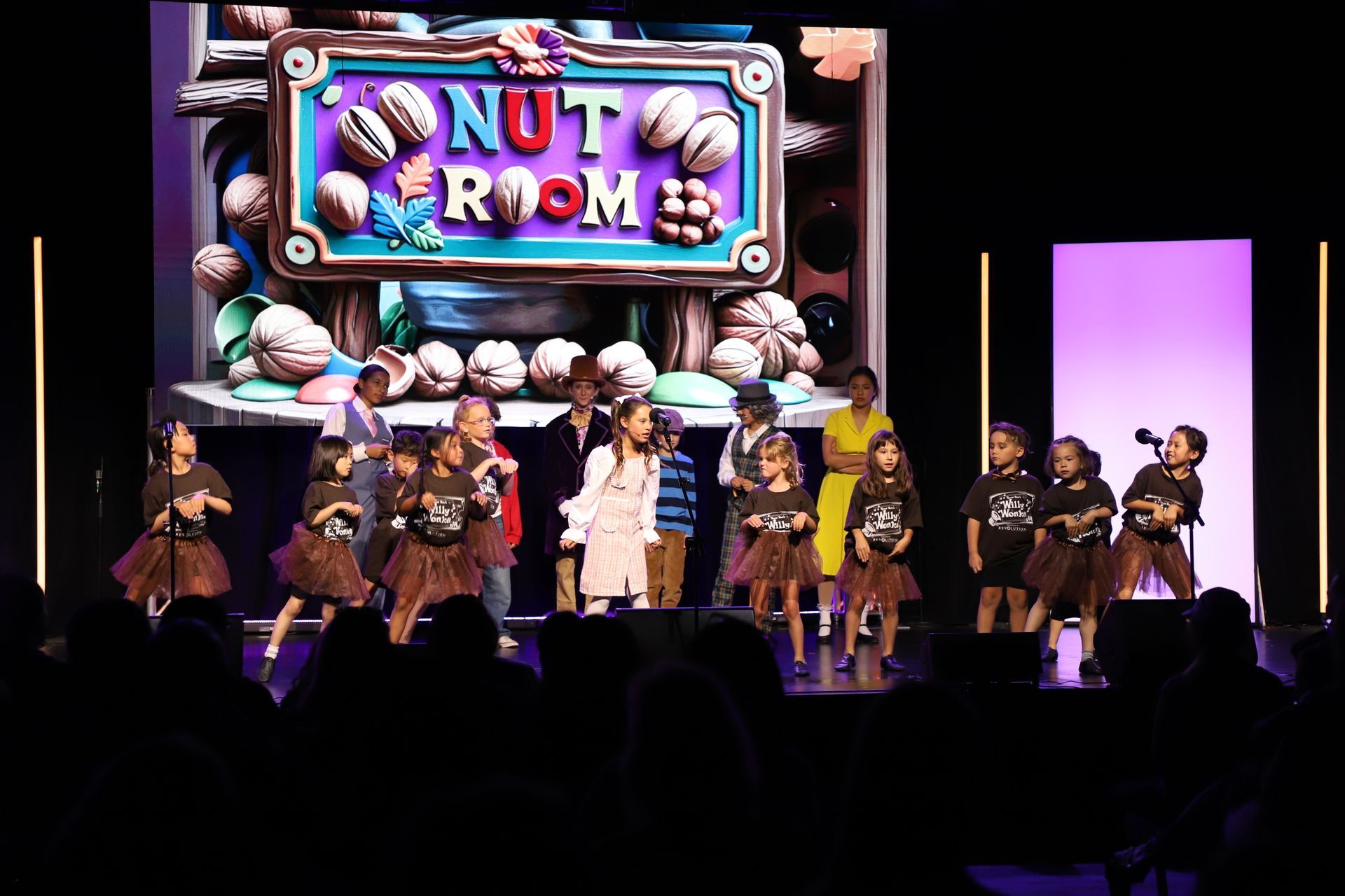 Children in costumes performing on stage under a colorful “Nut Room” sign for a theater performance.