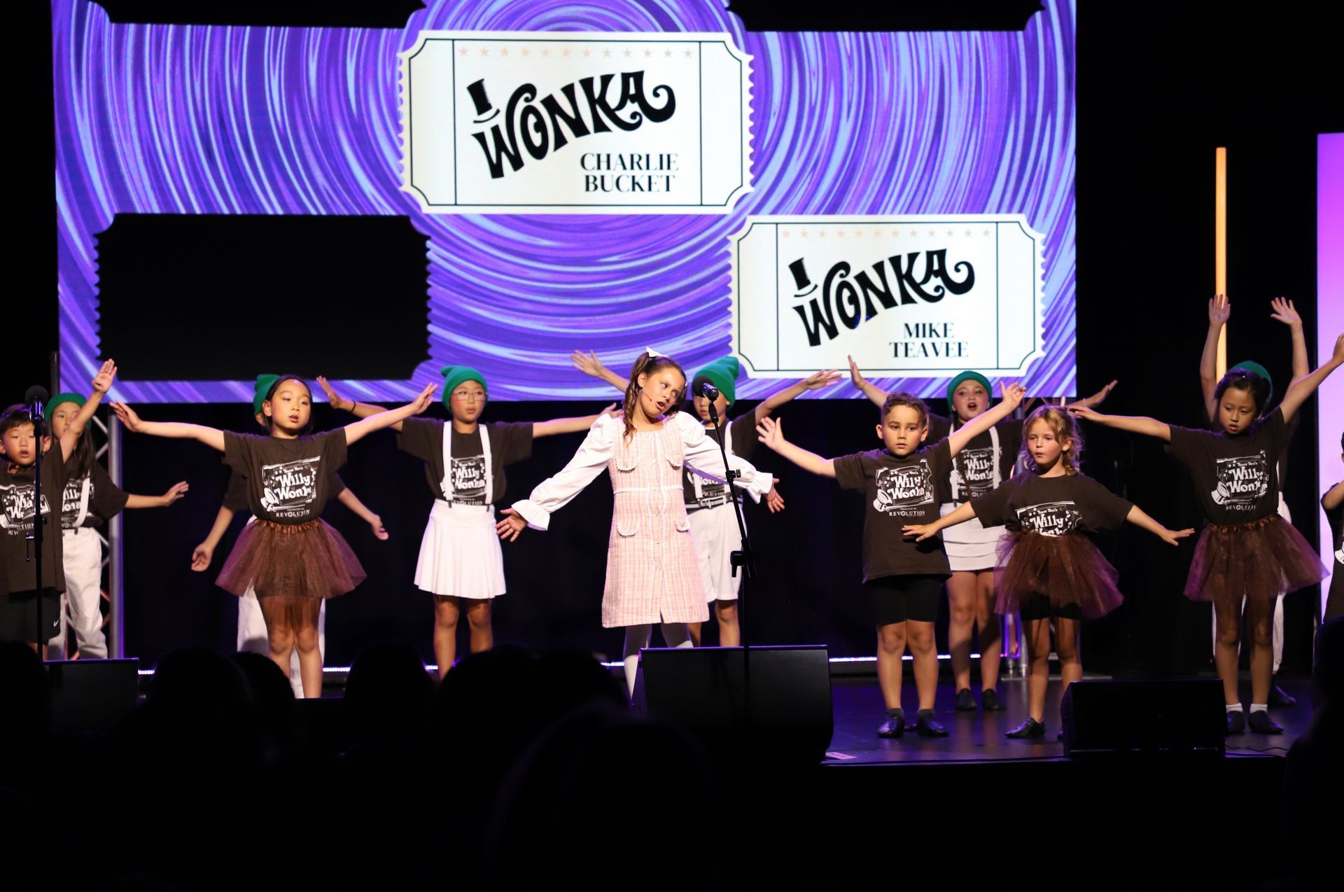 Performers on a Wonka-themed stage with purple backdrop, standing in a line and raising their arms for a theater performance.